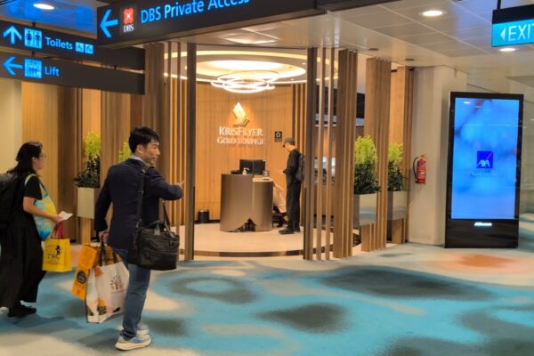 Approaching the KrisFlyer Gold Lounge entrance off the T2 concourse with the wood vestibule and lit signage above the reception podium.