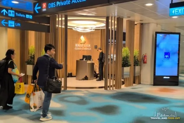 Approaching the KrisFlyer Gold Lounge entrance off the T2 concourse with the wood vestibule and lit signage above the reception podium.