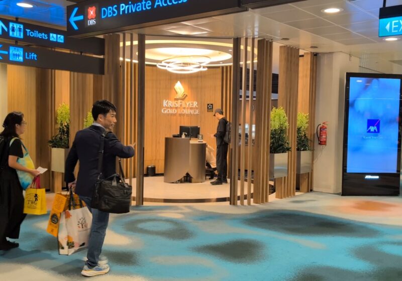 Approaching the KrisFlyer Gold Lounge entrance off the T2 concourse with the wood vestibule and lit signage above the reception podium.