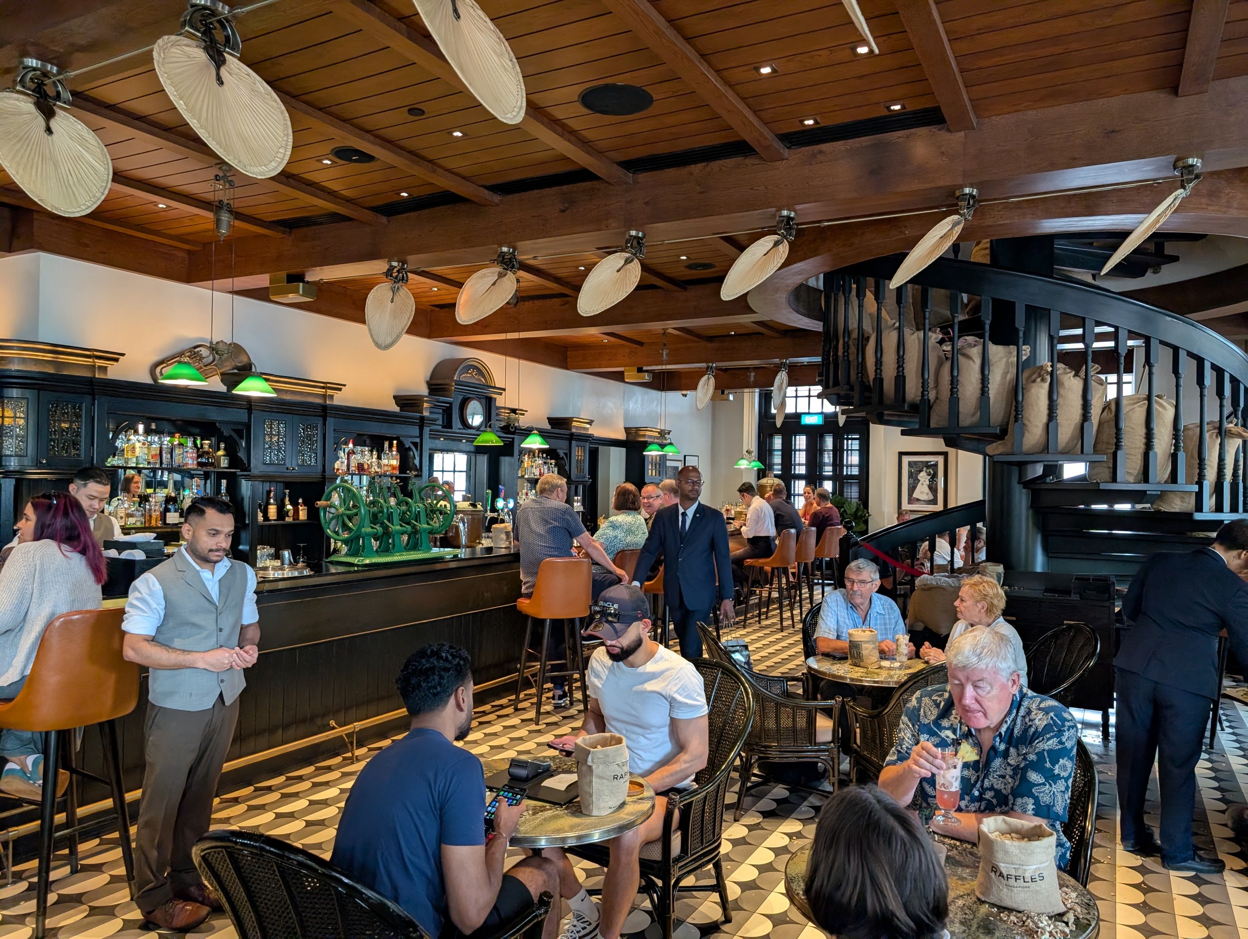 Long Bar interior at Raffles Hotel Singapore with palm leaf paddle ceiling fans, spiral staircase, and dark wood bar