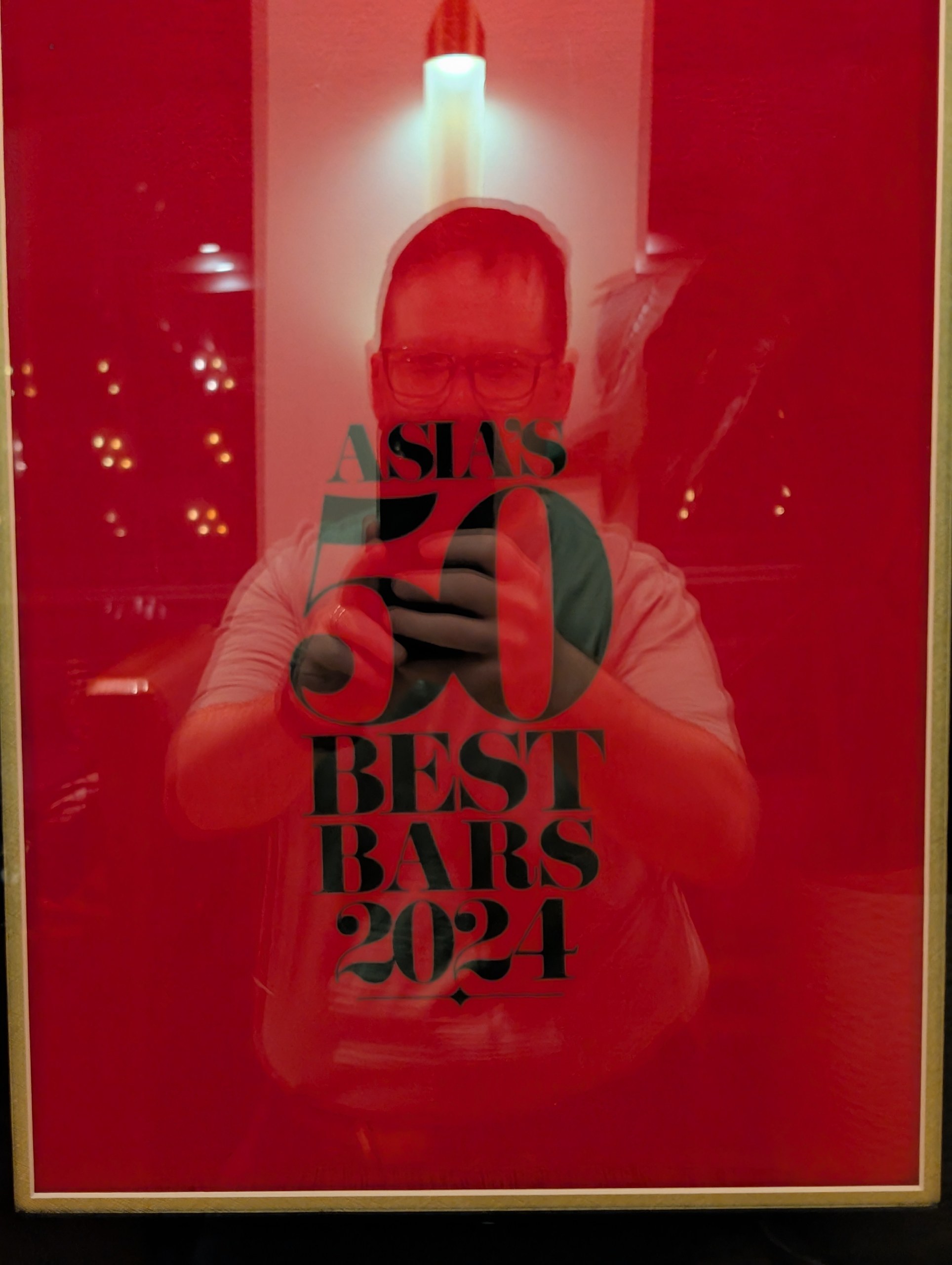 Asia's 50 Best Bars 2024 poster behind glass at the entrance to Origin Grill and Bar, with the photographer's reflection in the glass.