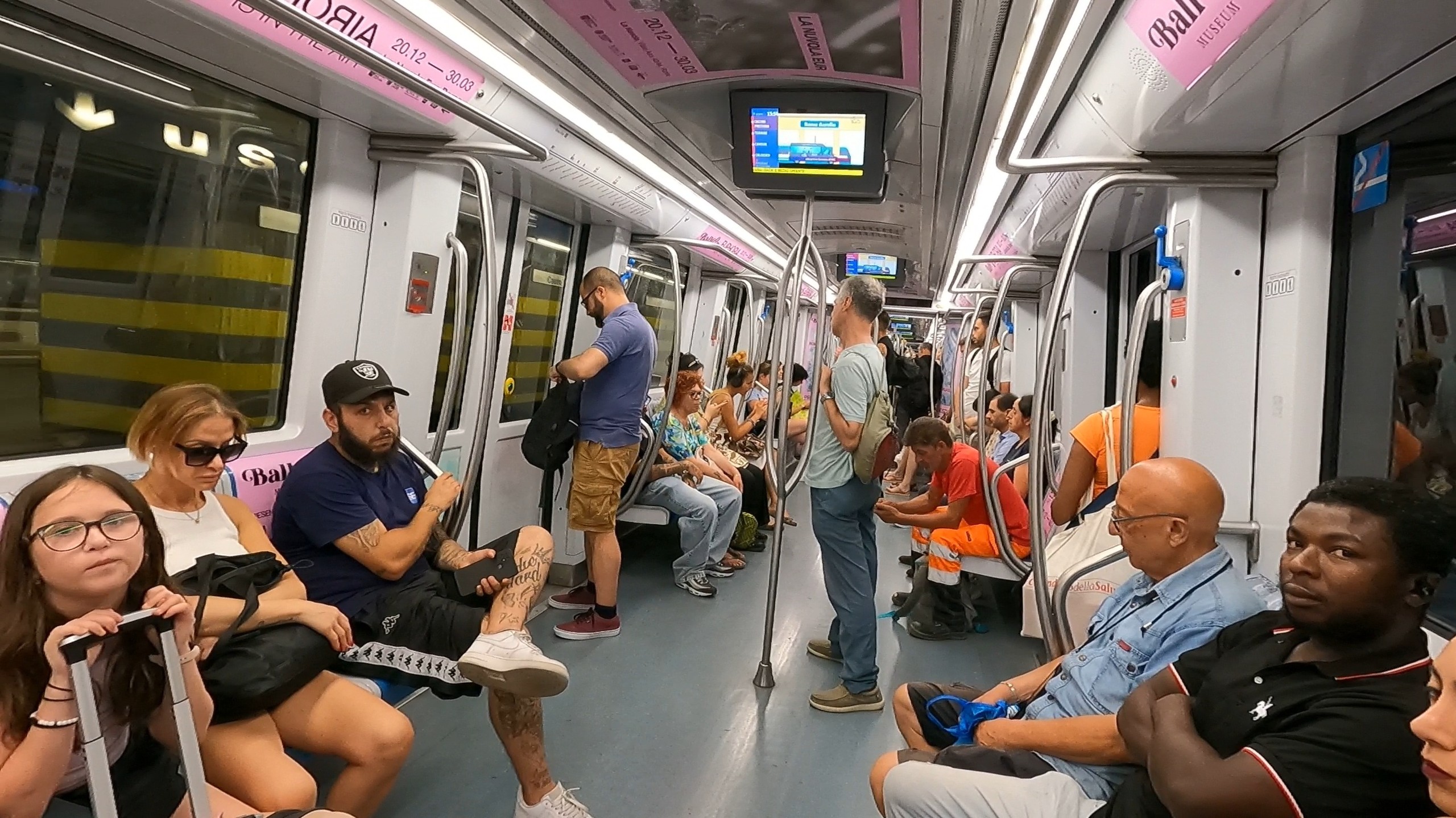 Inside a Rome metro car during a daytime run with passengers seated and standing in the aisle