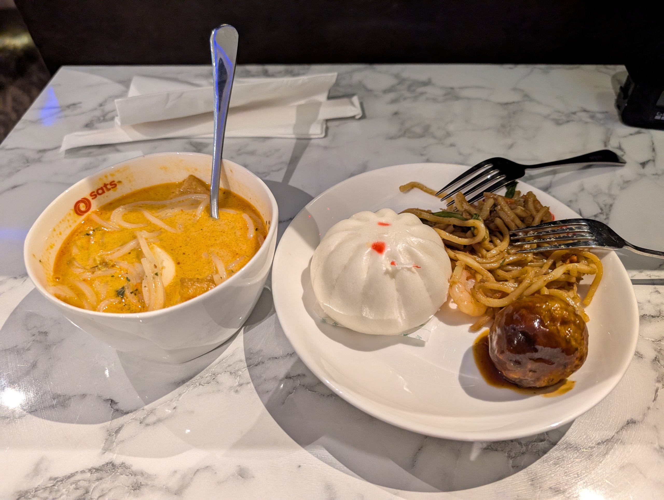 A bowl of signature laksa, a steamed bao bun, stir fried noodles, and a meatball at the SATS Premier Lounge Changi T2