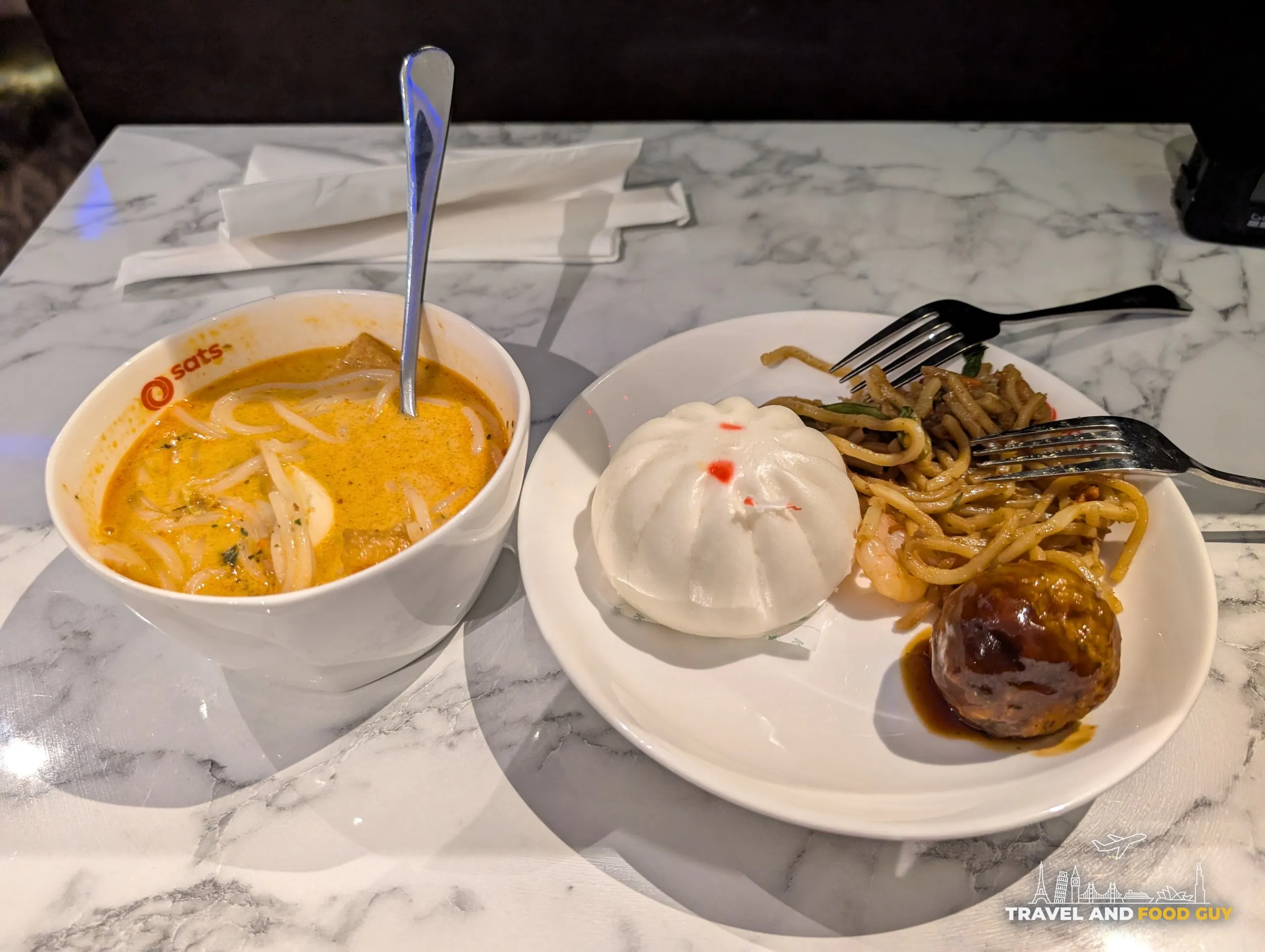 A SATS-branded bowl of signature laksa next to a plate with a bao bun, mee goreng noodles, and a glazed meatball at the SATS Premier Lounge Changi T2.