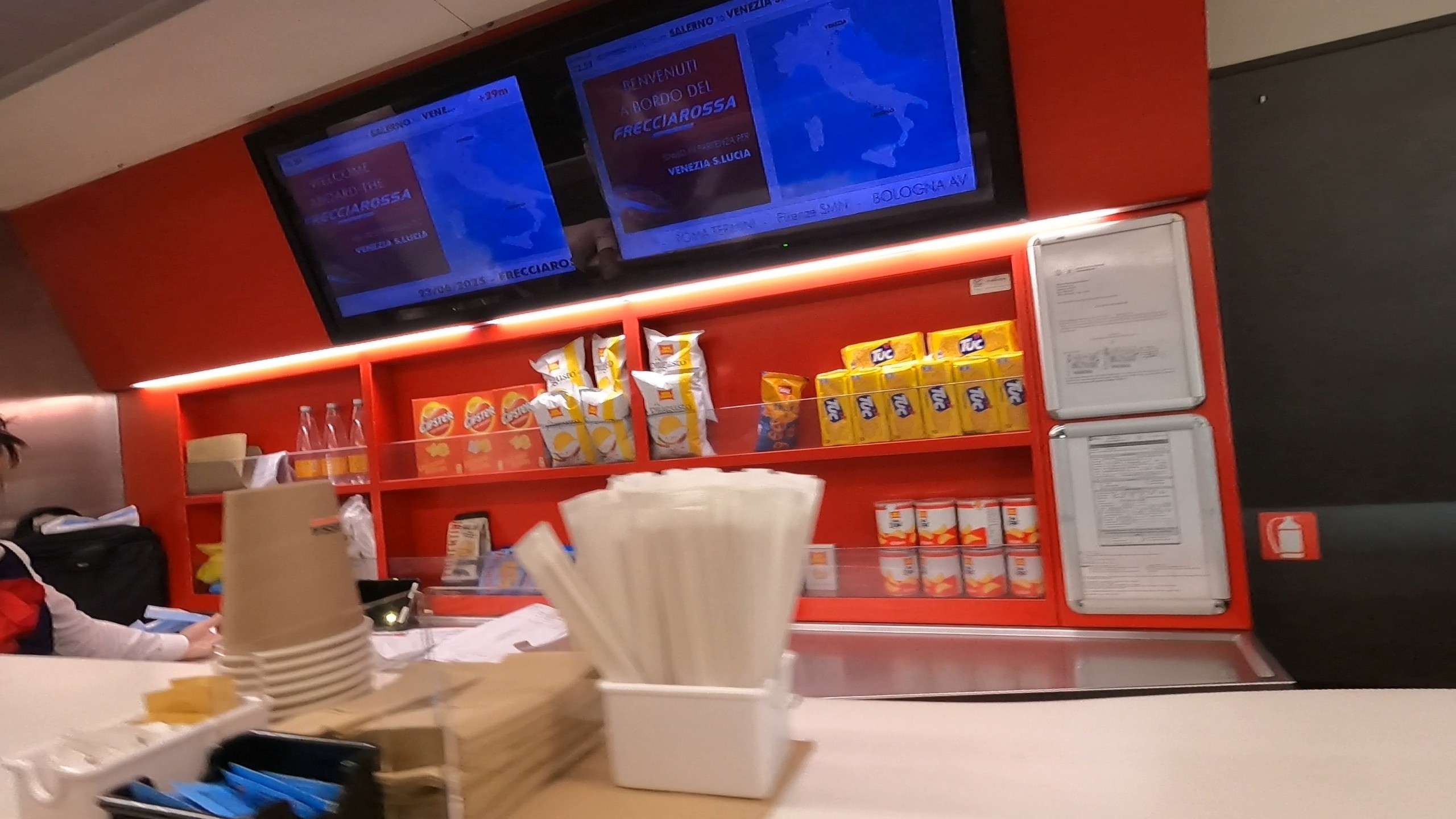 Onboard Trenitalia Frecciarossa snack service with packaged crackers and drinks beneath the welcome monitor
