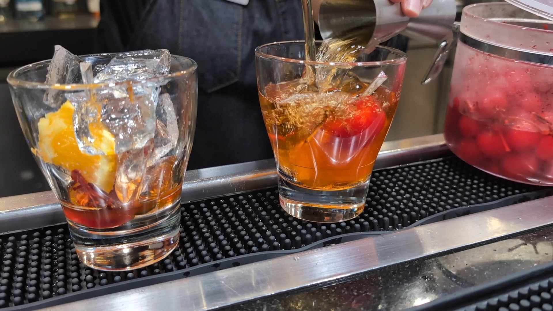 Two Old Fashioneds being built at the bar at the United Club at IAH Gate C33, with orange peel and ice in glass tumblers and a jar of cherries on the rail.