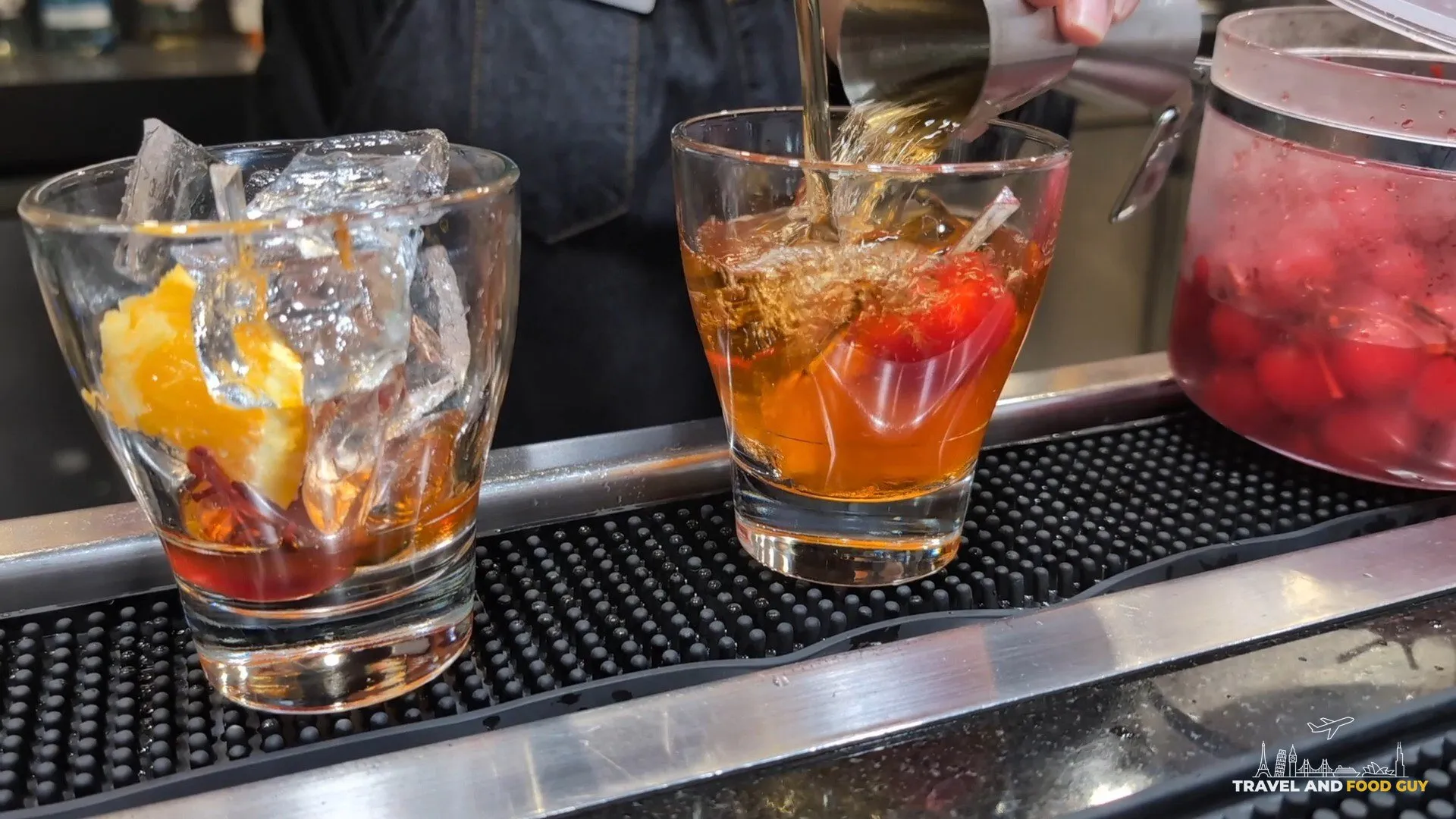Two Old Fashioneds being built at the bar at the United Club at IAH Gate C33, with orange peel and ice in glass tumblers and a jar of cherries on the rail.