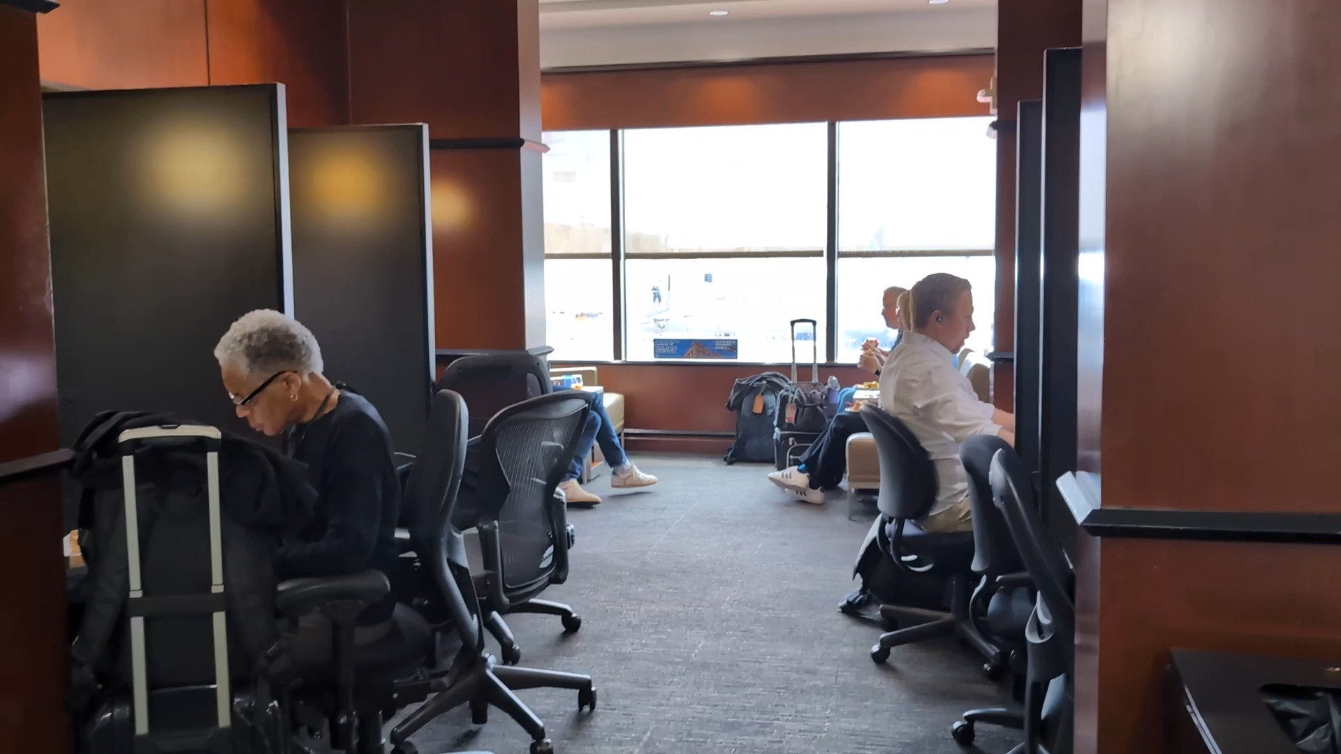 Window-side workspace at the United Club at IAH Gate C33 with a partial tarmac view, two travelers at task chairs in front of monitors.