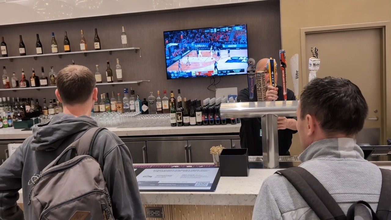 Full bar at the United Club at ORD Gate B18, with bottles of wine on the back shelf, a TV showing a basketball game, and two travelers on bar stools.