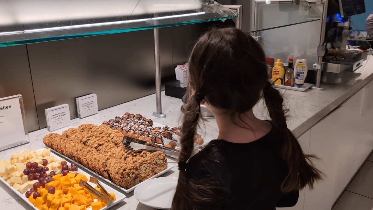 Trays of cookies, mini cheese cubes, and grapes on the food counter at the United Club at ORD Gate B18, with my daughter Emily reaching in for a snack.