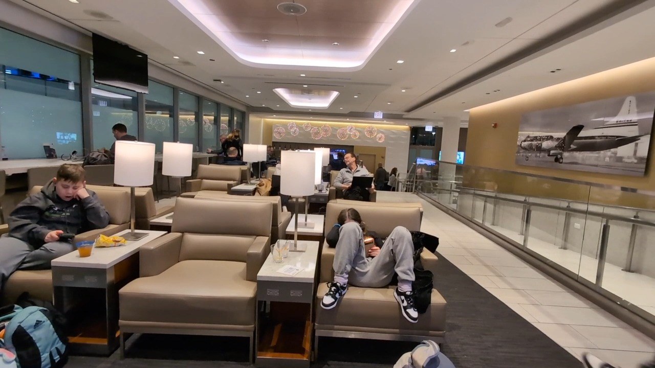 My son Josh on a club chair with his phone and a yellow drink, and my daughter Emily reclined on the couch across from him at the United Club at ORD Gate B18.