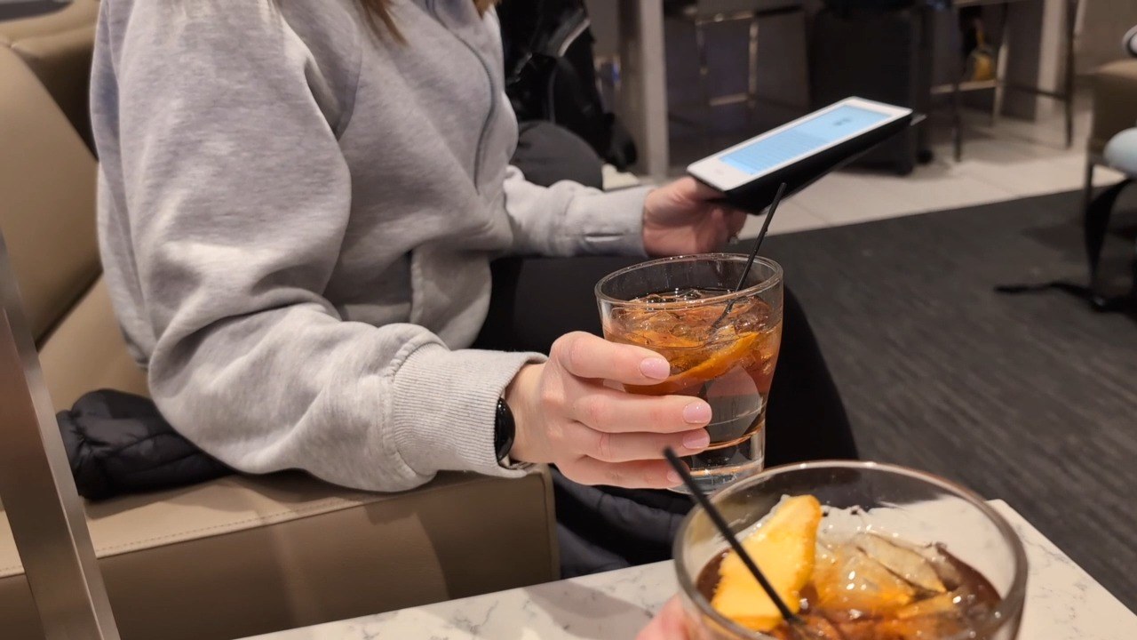 Old Fashioned in a glass with orange peel held by my wife Vika at the United Club at ORD Gate B18, with a tablet open in her other hand.