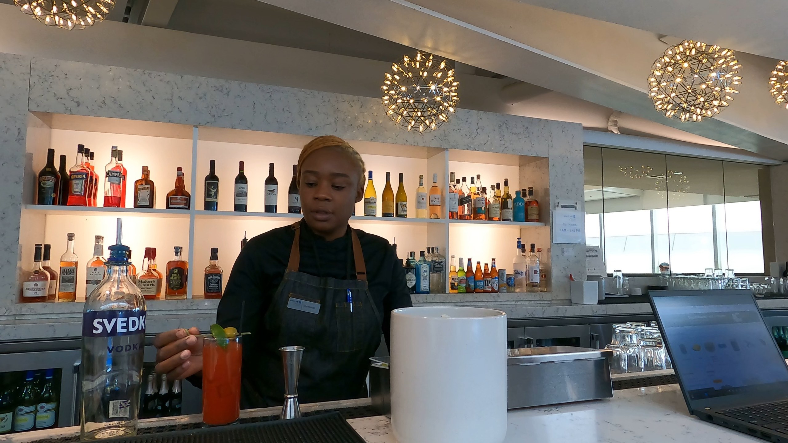 Bartender at the bar at the United Club at ORD Gate B6 making a Bloody Mary, with bottles of Svedka and other spirits on the back wall and the starburst chandeliers behind.