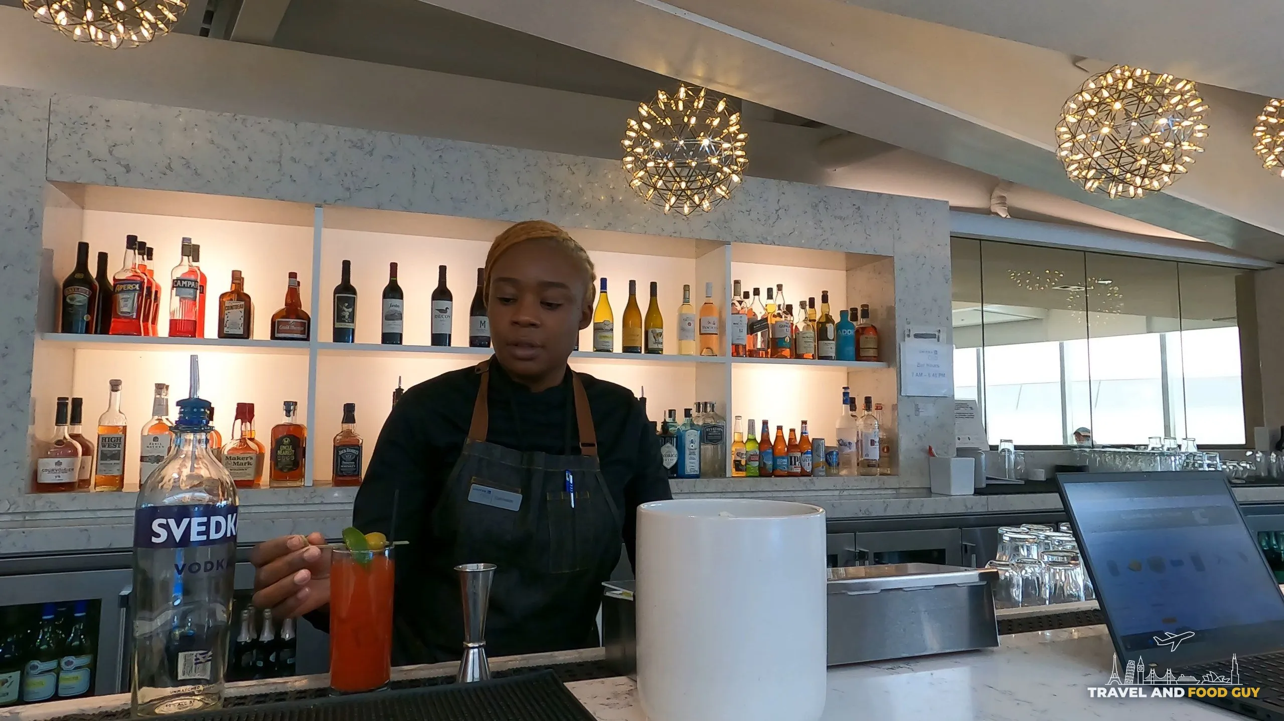 Bartender at the bar at the United Club at ORD Gate B6 making a Bloody Mary, with bottles of Svedka and other spirits on the back wall and the starburst chandeliers behind.