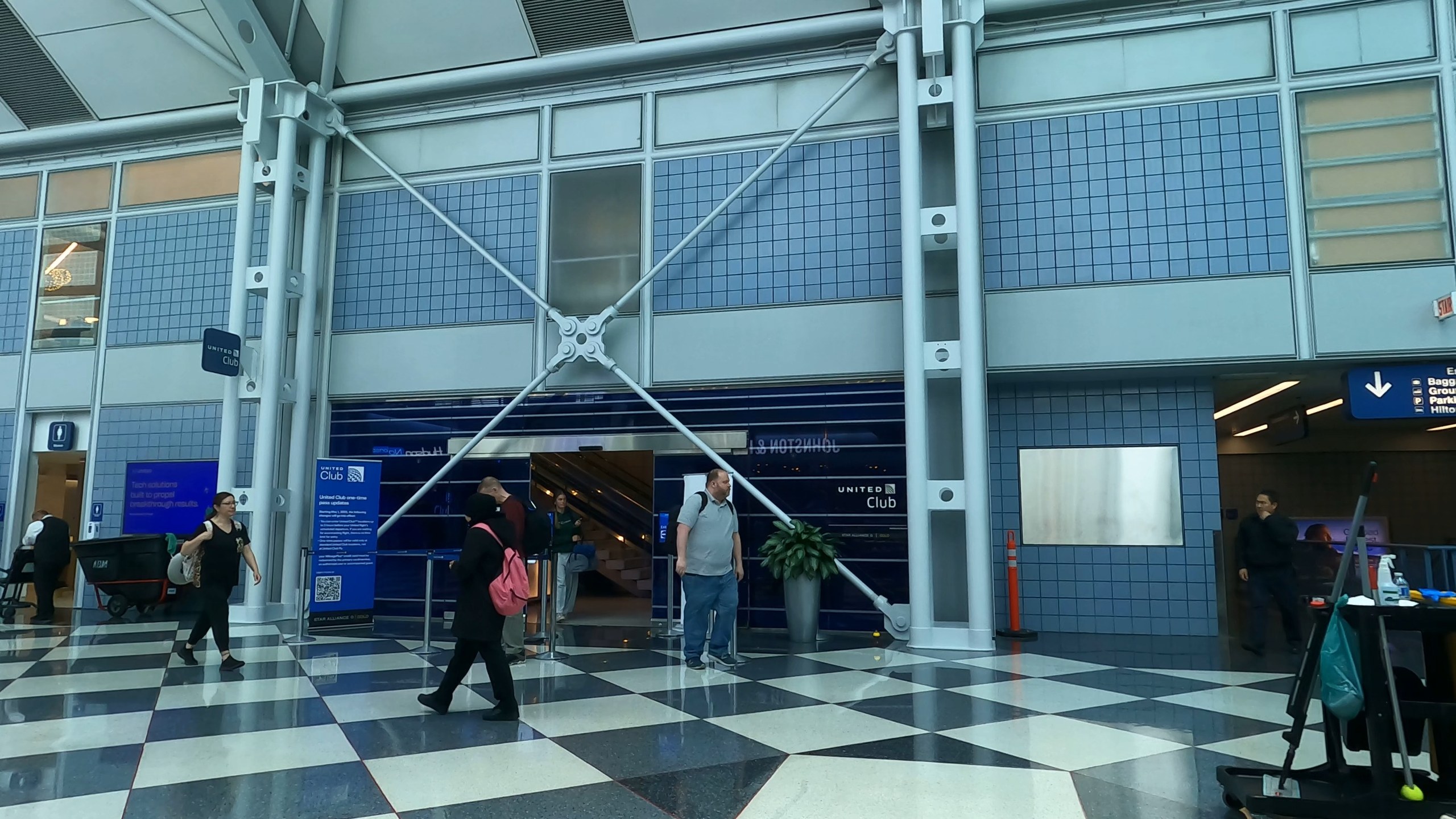 Exterior view of the United Club at ORD Gate B6 from the concourse below, with the black-and-white checkerboard floor in the foreground and the United Club entrance flanked by structural braces.