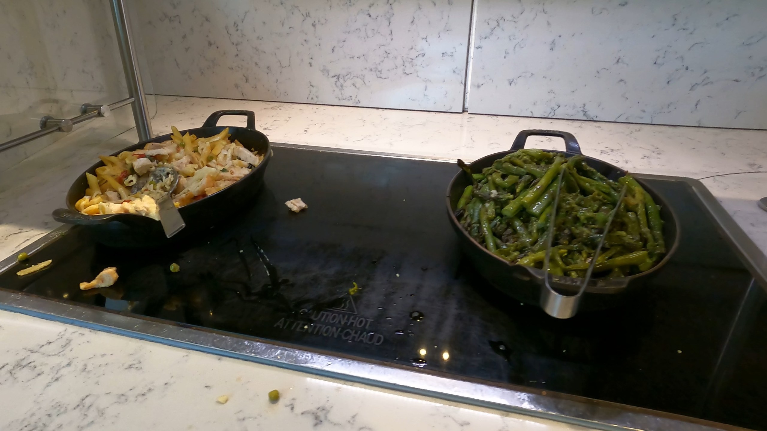Two cast-iron skillets of hot food, penne pasta with chicken on the left and seasoned green beans on the right, on the warming plate at the United Club at ORD Gate B6.