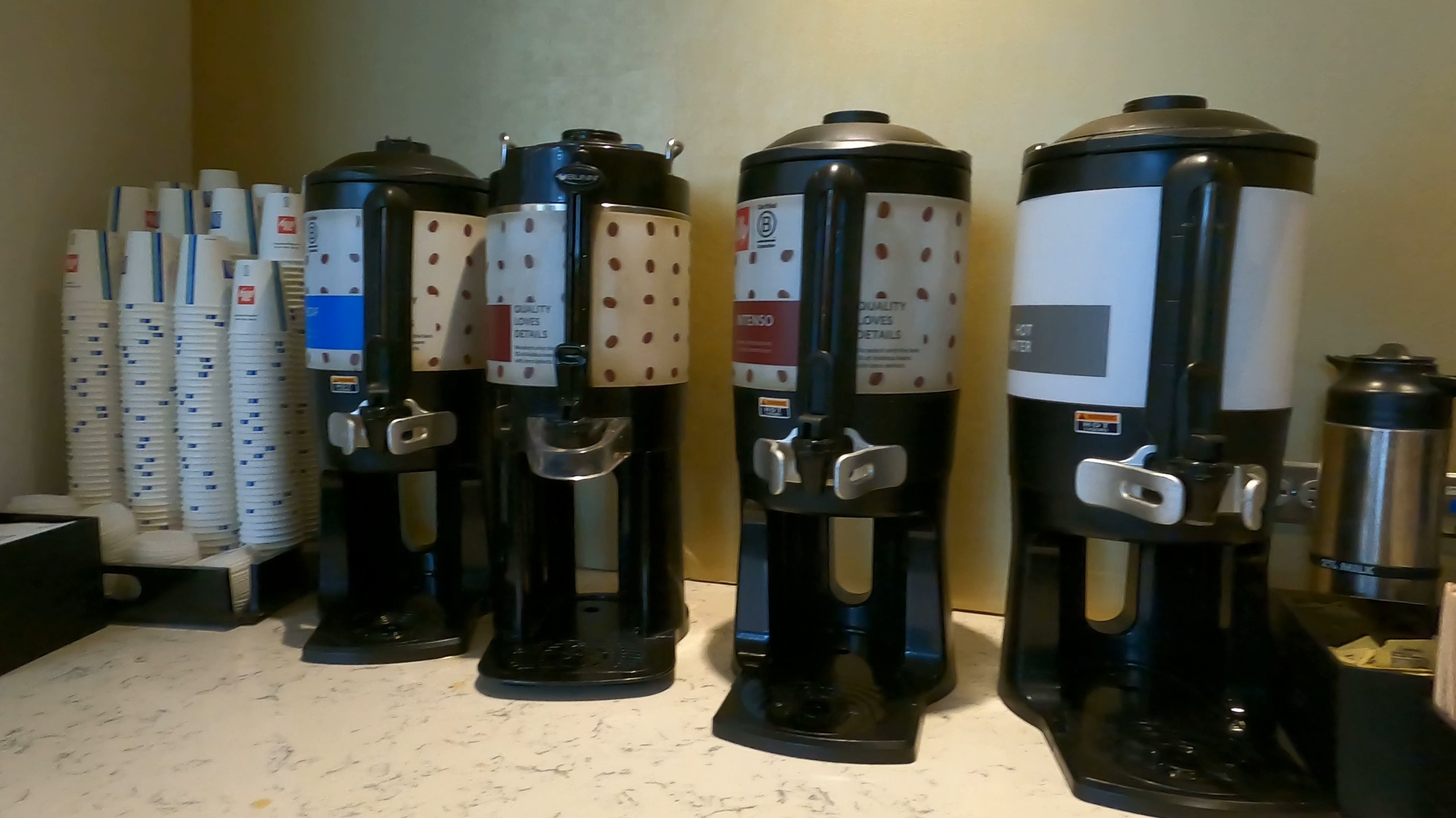 Bank of four Illy coffee dispensers at the United Club at ORD Gate B6, marked decaf, regular, intenso, and hot water, with a tower of branded paper cups on the left.