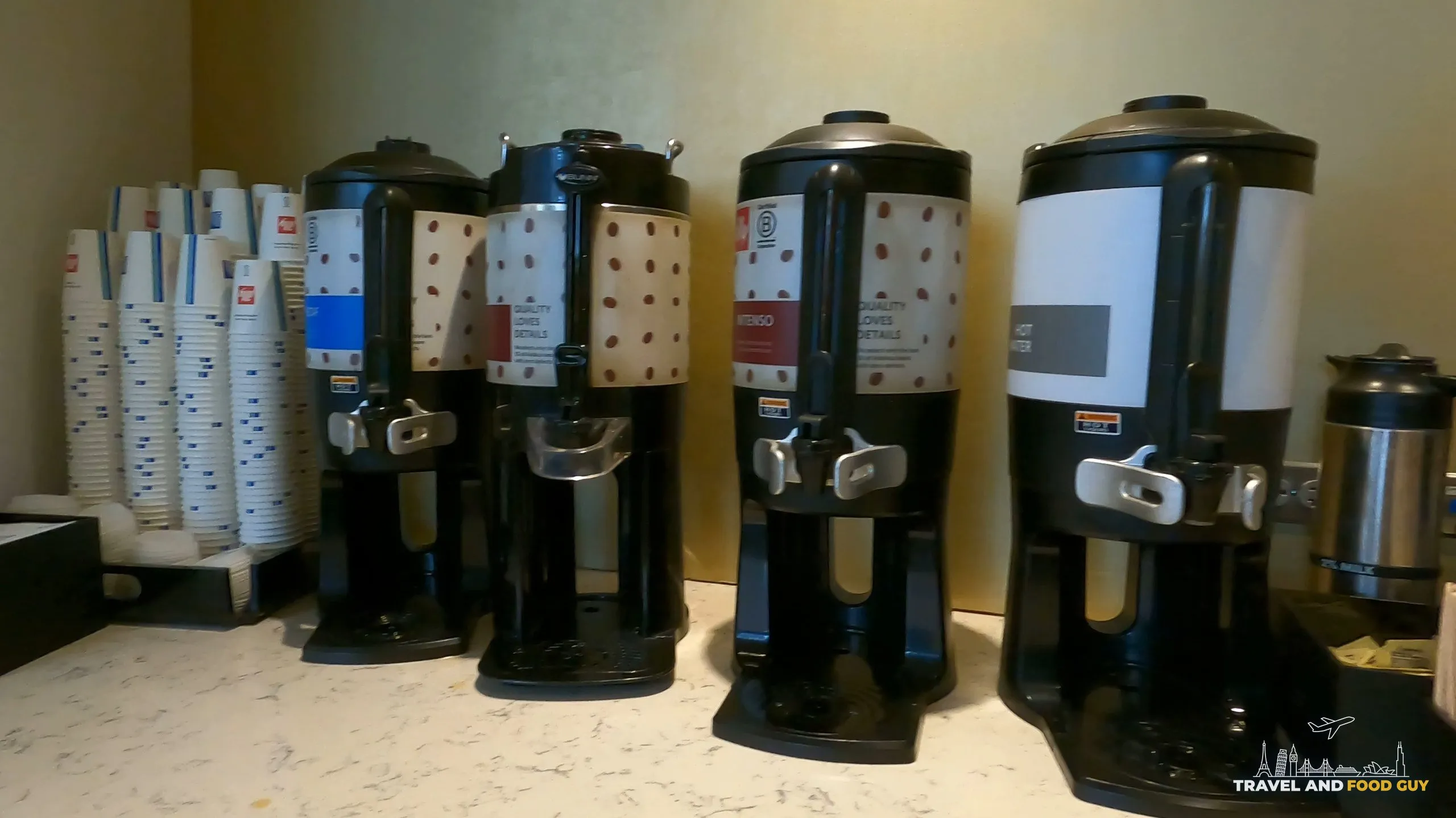 Bank of four Illy coffee dispensers at the United Club at ORD Gate B6, marked decaf, regular, intenso, and hot water, with a tower of branded paper cups on the left.