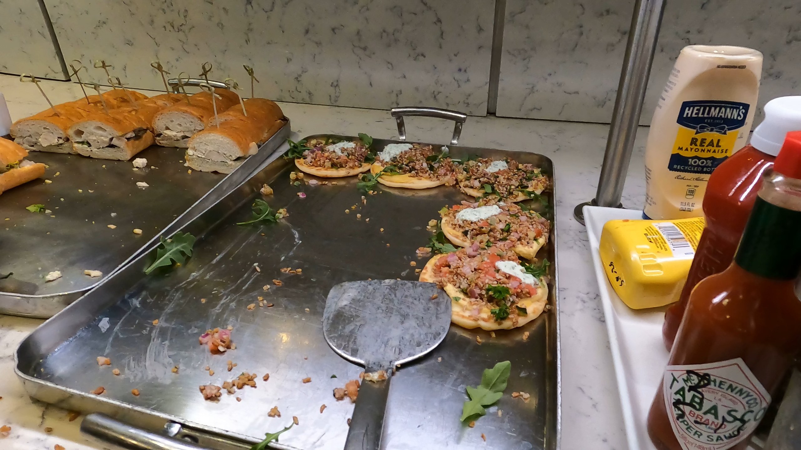 Hot food station at the United Club at ORD Gate B6 with mini personal flatbreads, sliced baguette, and a tray of mayonnaise, mustard, and Tabasco sauce.