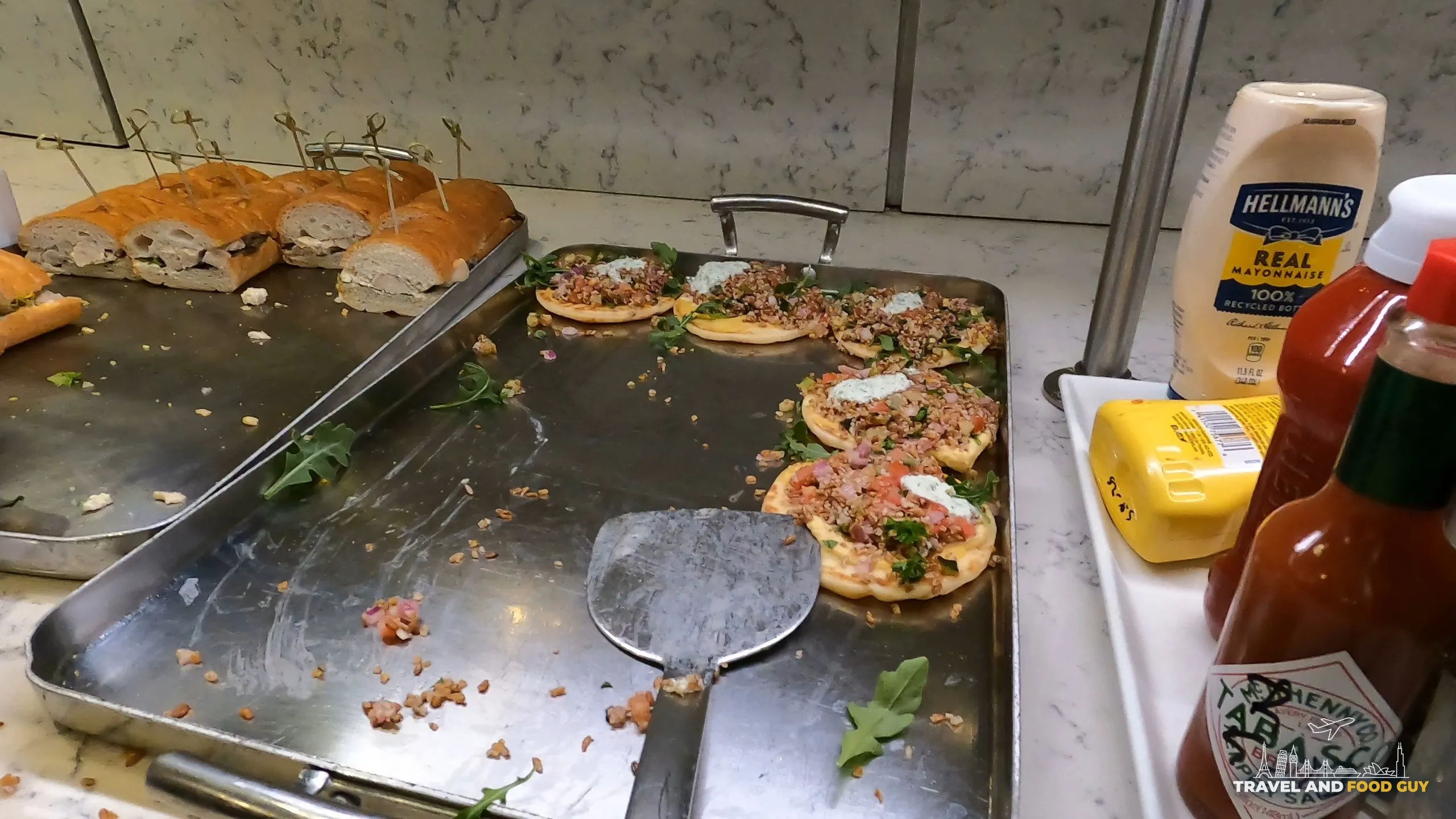 Hot food station at the United Club at ORD Gate B6 with mini personal flatbreads, sliced baguette, and a tray of mayonnaise, mustard, and Tabasco sauce.