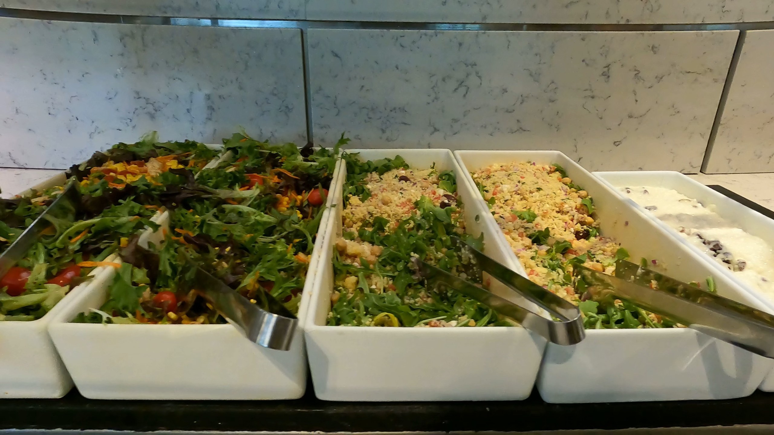 Salad bar at the United Club at ORD Gate B6 with mixed greens, two grain salads, a Greek-style salad, and a yogurt or feta-and-olive option in white serving dishes.