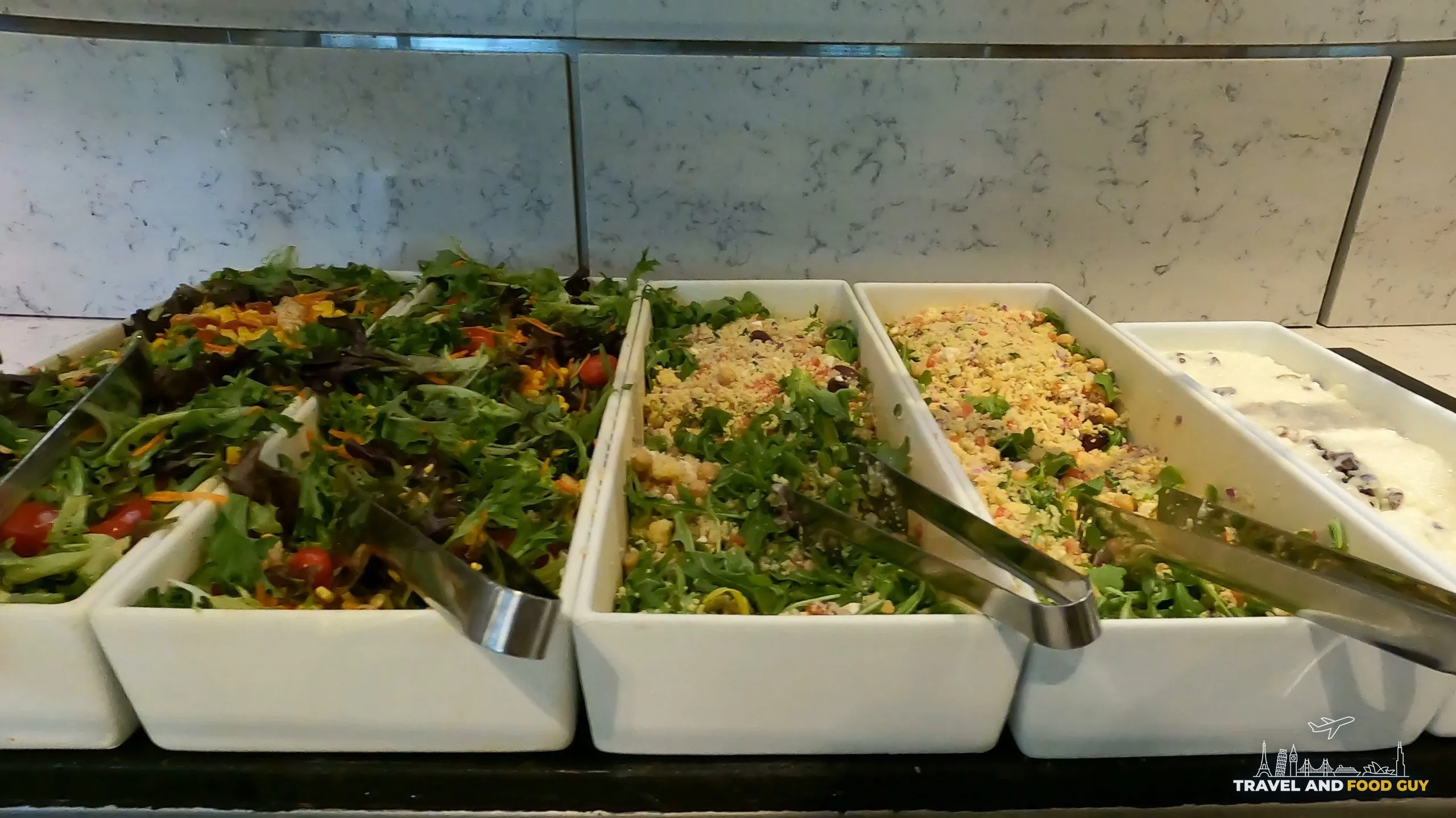 Salad bar at the United Club at ORD Gate B6 with mixed greens, two grain salads, a Greek-style salad, and a yogurt or feta-and-olive option in white serving dishes.