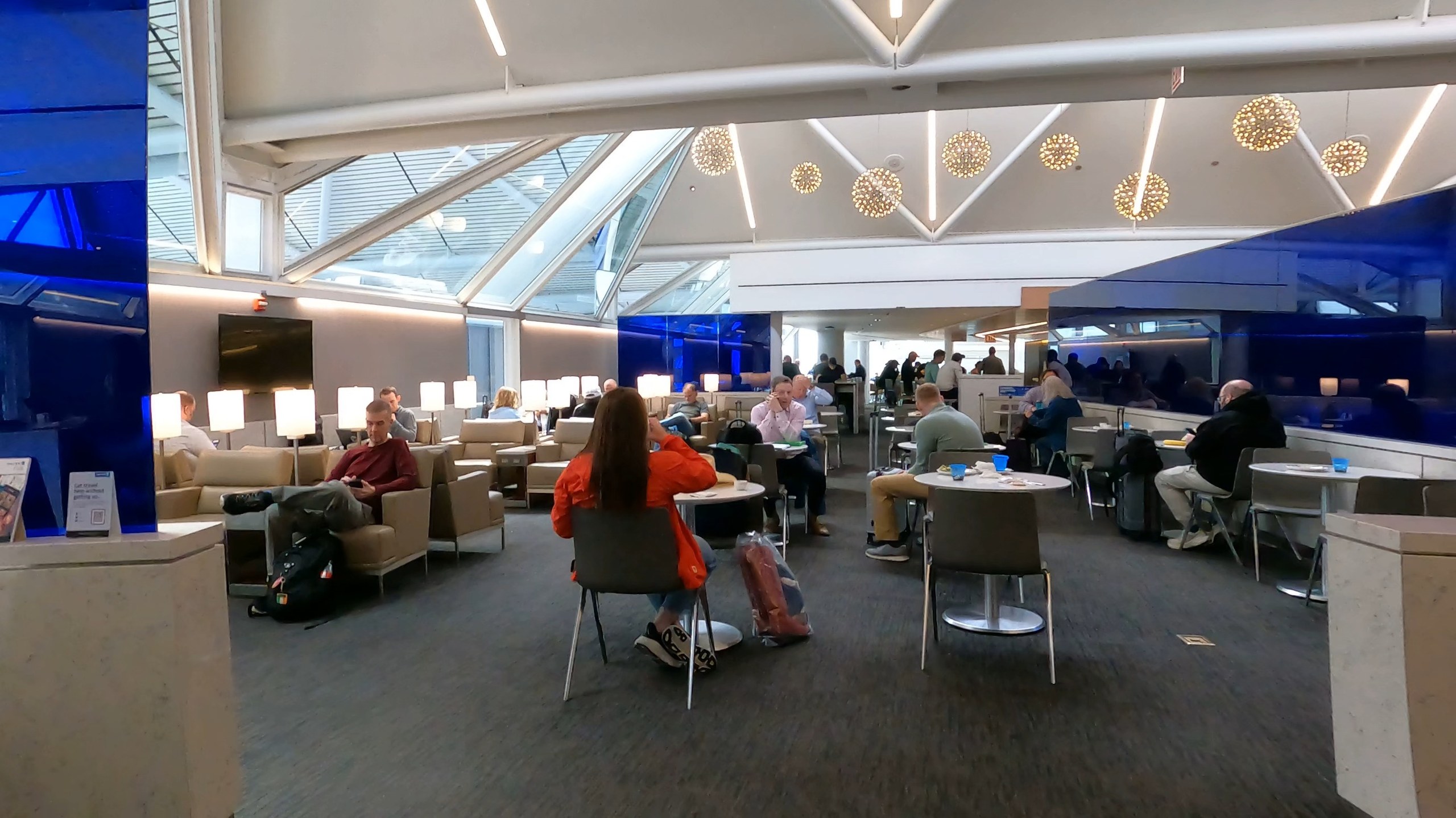 Wide view of the seating at the United Club at ORD Gate B6 under the skylit ceiling, with starburst chandeliers, leather chairs, and travelers working at small round tables.