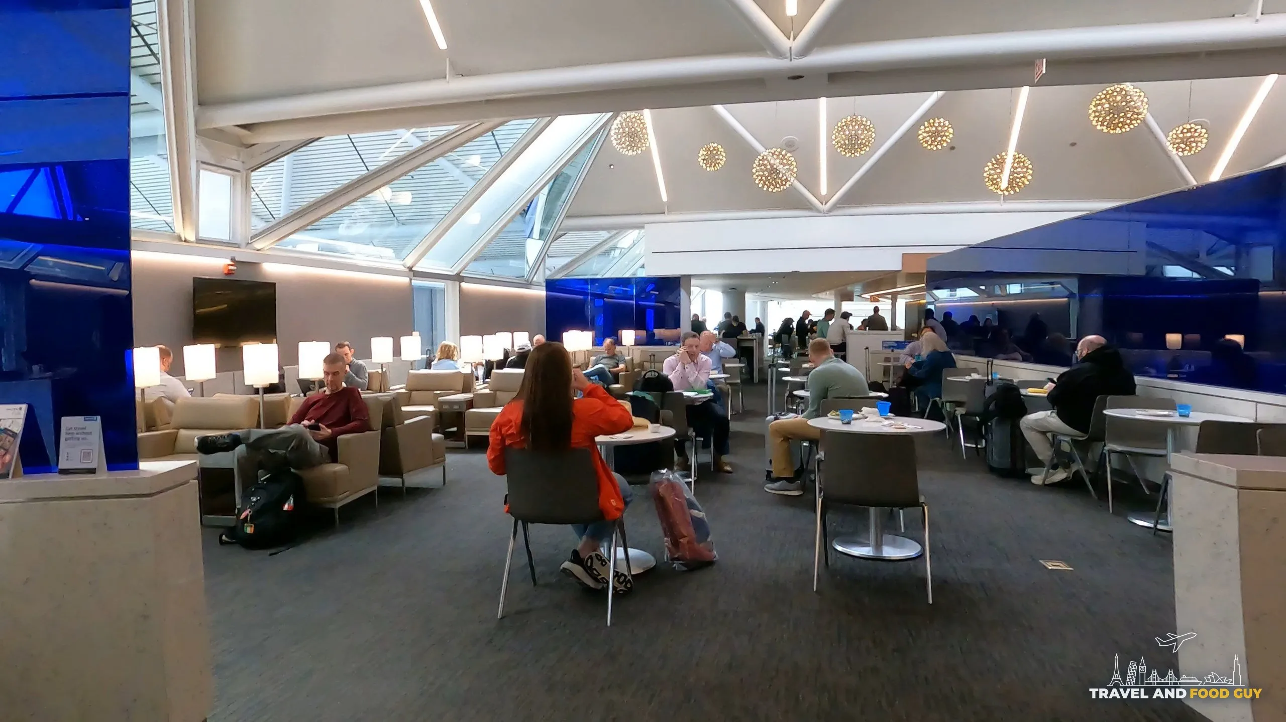 Wide view of the seating at the United Club at ORD Gate B6 under the skylit ceiling, with starburst chandeliers, leather chairs, and travelers working at small round tables.