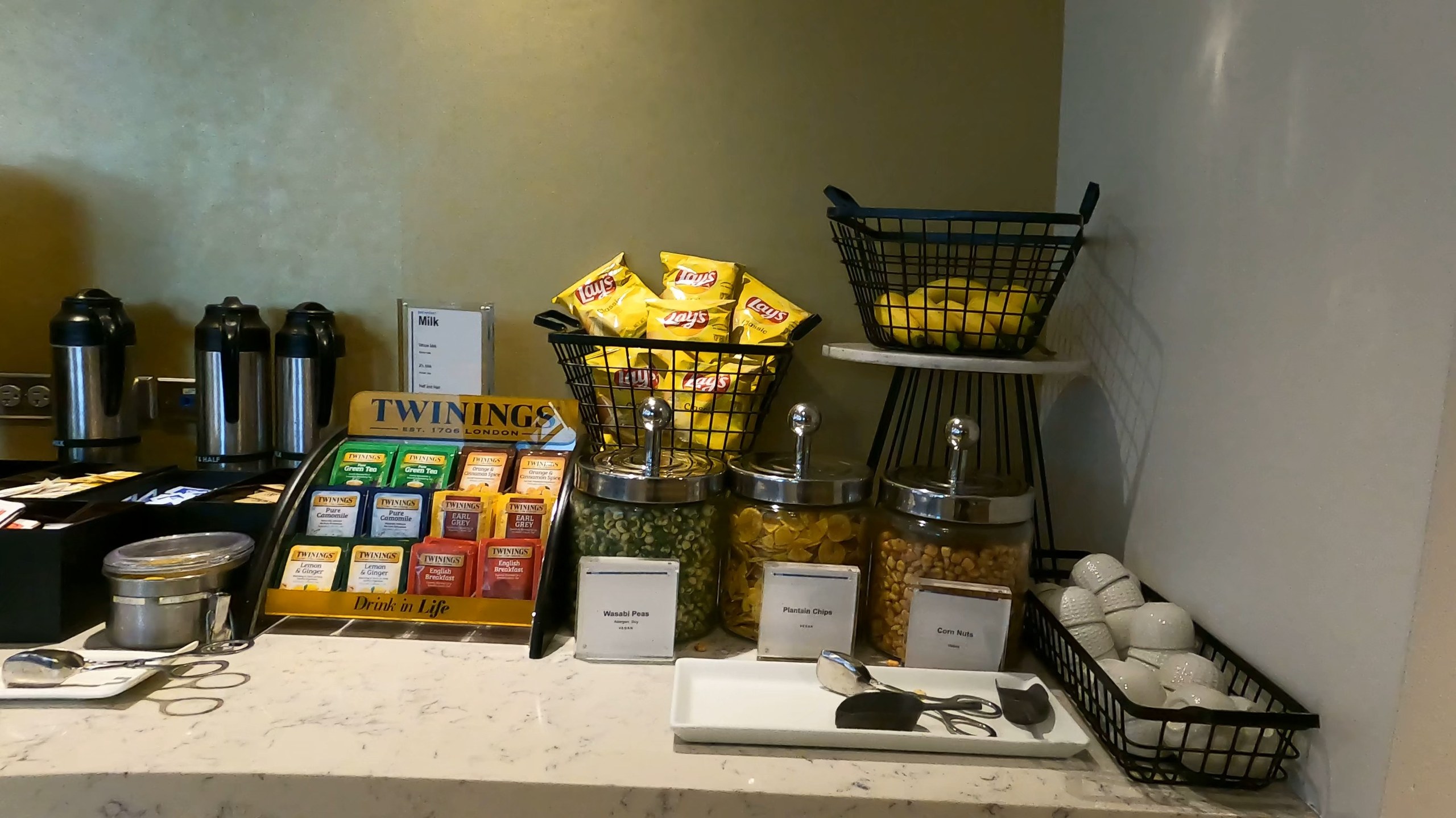 Snack station at the United Club at ORD Gate B6 with Twinings tea, coffee thermoses, baskets of Lay's chips, and three apothecary jars of mixed snacks.