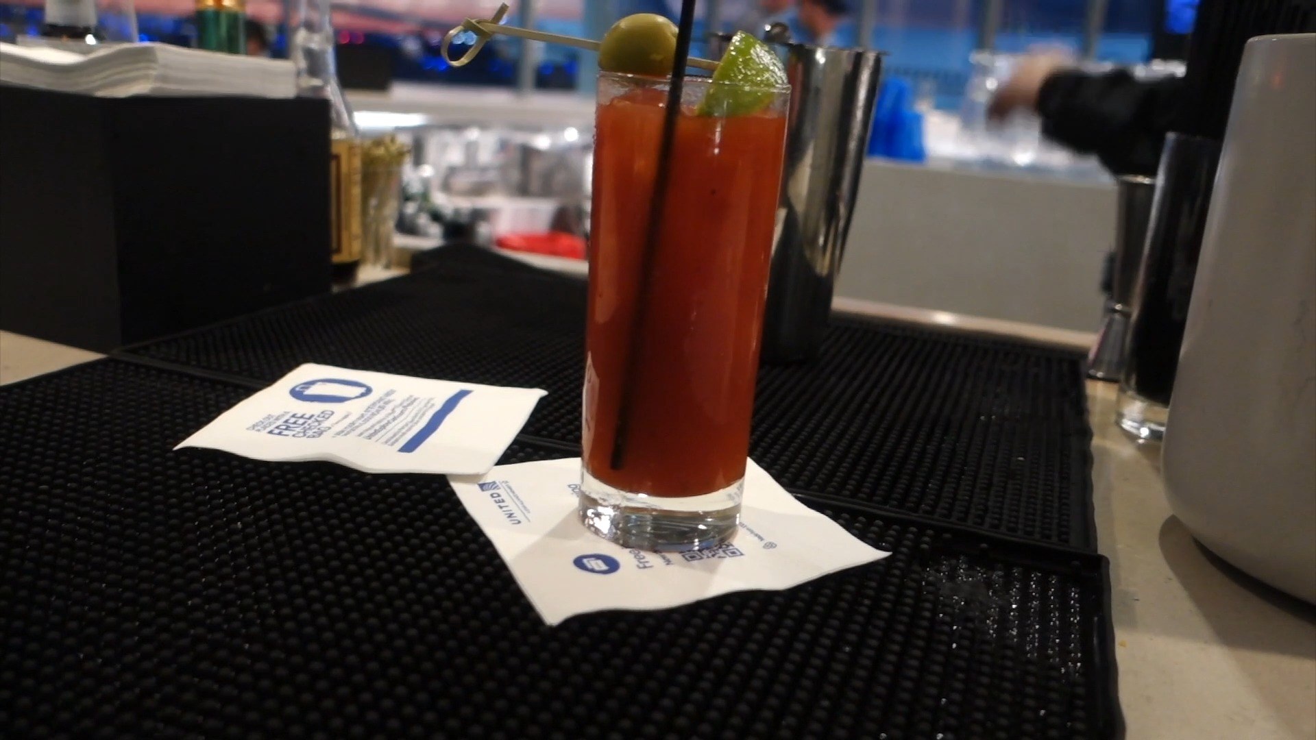 A Bloody Mary in a tall highball with green olive, lime wedge, and a straw on the black bar mat at the United Club at ORD Concourse C, on a United-branded napkin.