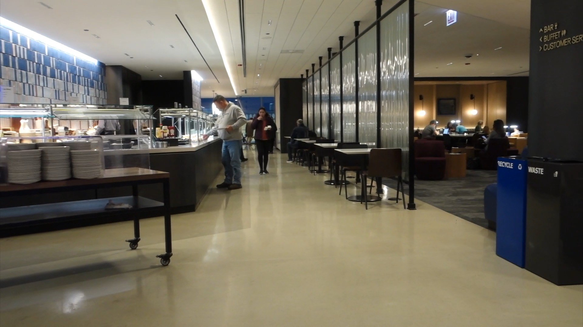 The buffet line at the United Club at ORD Concourse C with hot-food chafing dishes, plates stacked on a wooden cart, communal seating beyond, and a fluted-glass divider on the right.