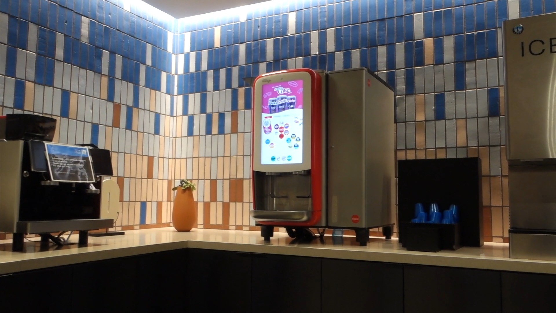 Coca-Cola Freestyle machine and an automatic espresso machine on the tiled counter at the United Club at ORD Concourse C, with an orange vase and orchid in between.