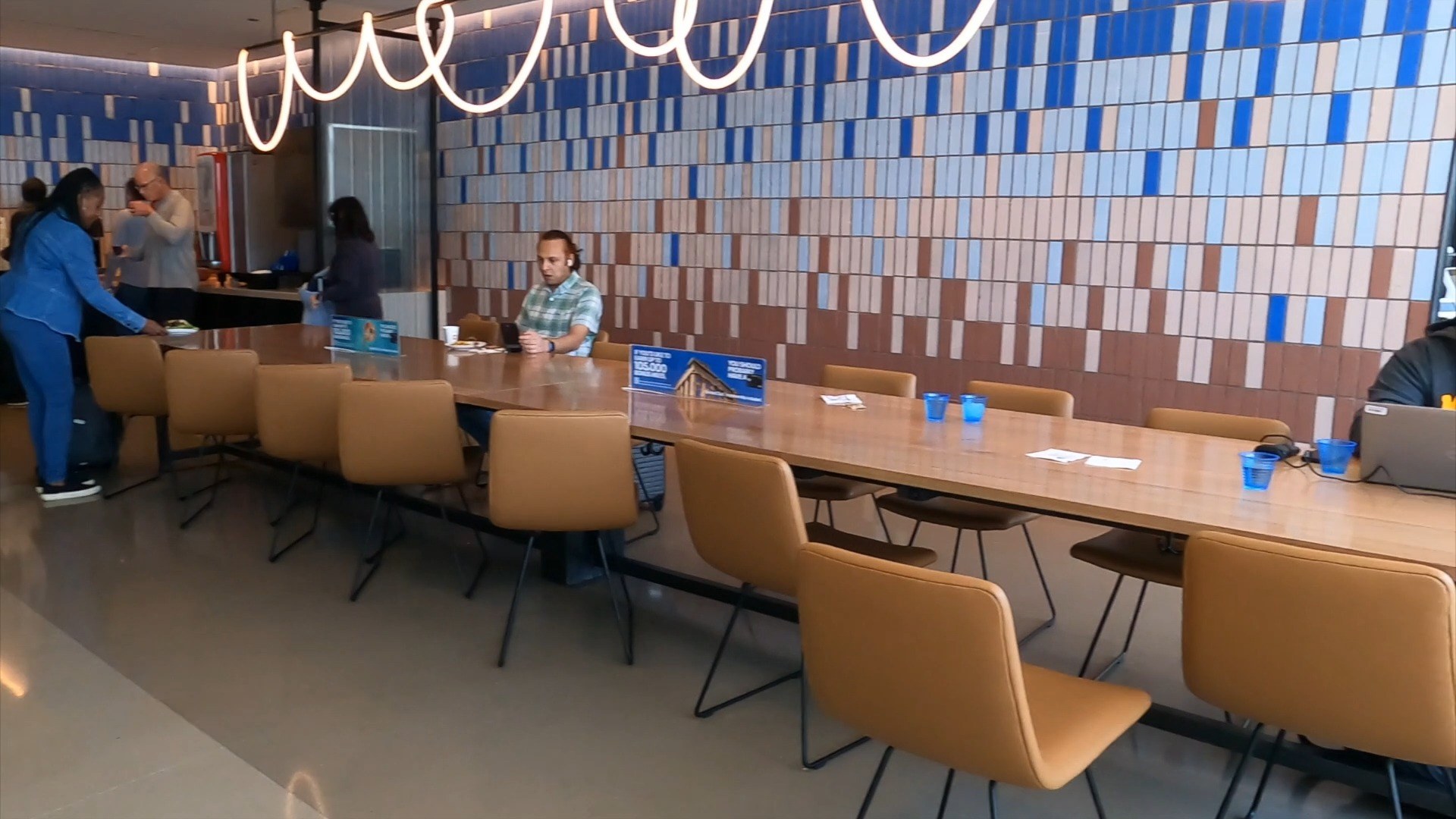 Long wooden communal table at the United Club at ORD Concourse C with curved pendant lamps overhead and the blue, cream, and orange tile mosaic wall behind.