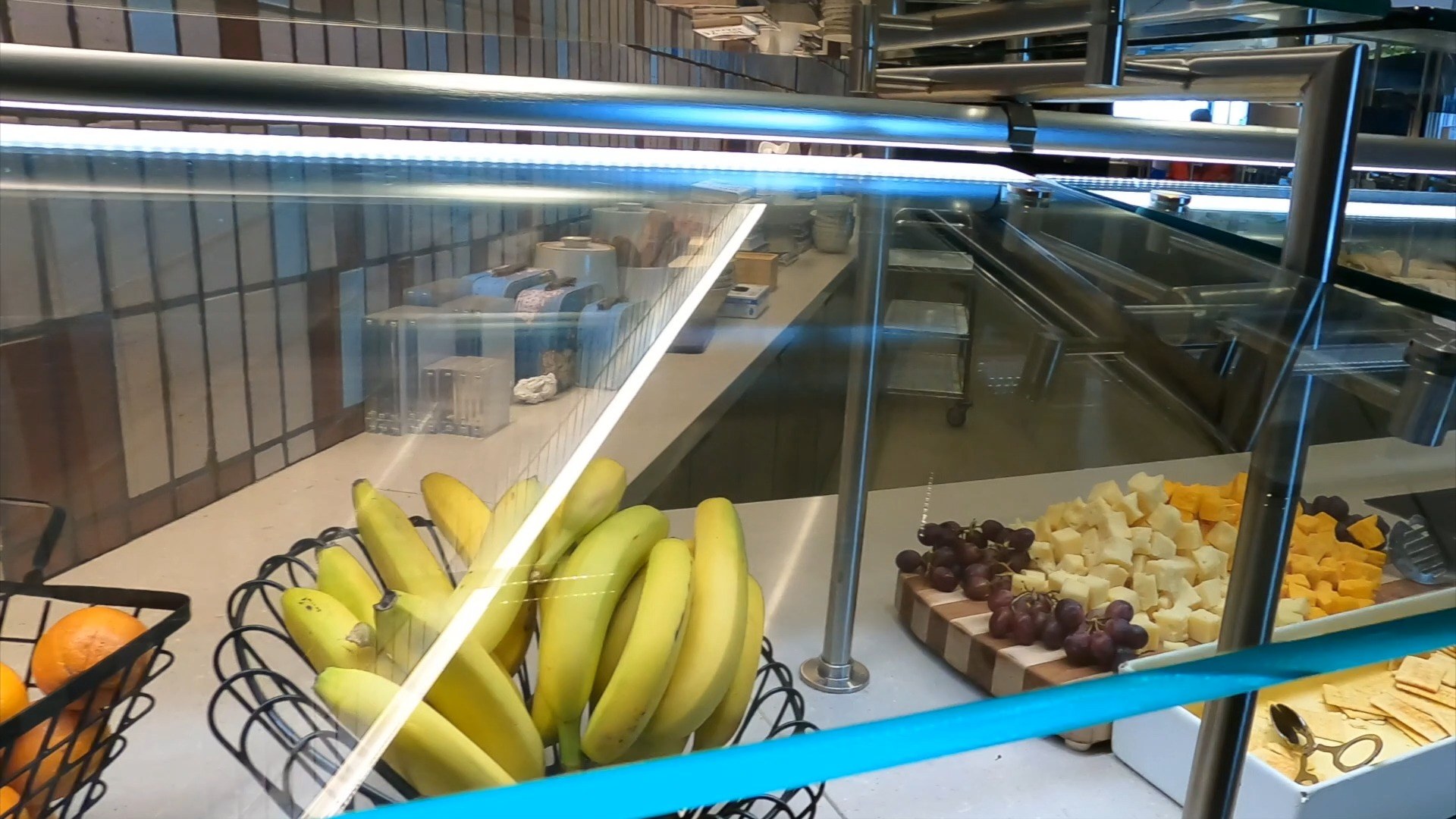 Fruit and cheese display at the United Club at ORD Concourse C with bananas in a wire basket, cheese cubes and grapes on a wooden cutting board, and oranges in another basket.
