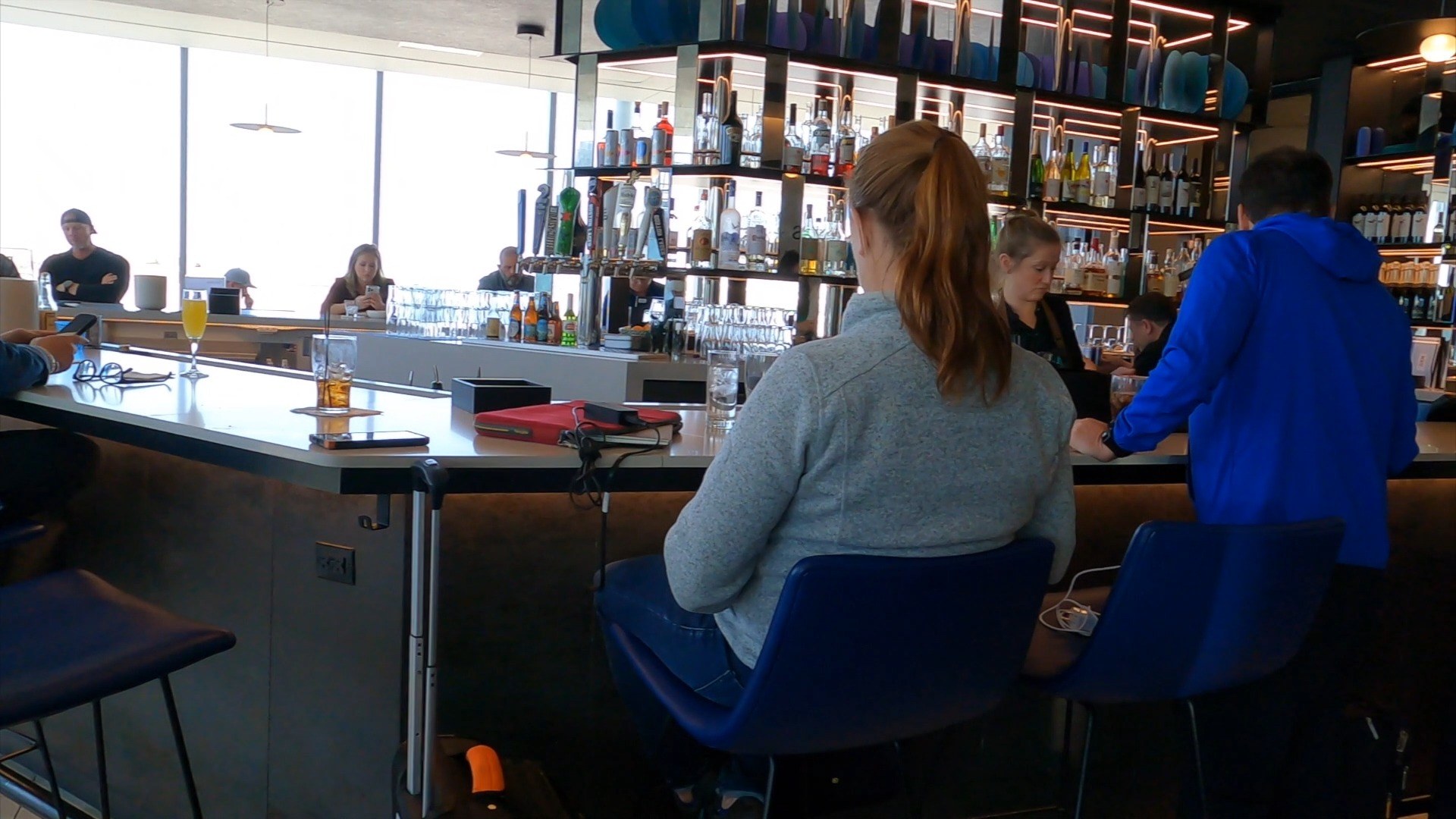The full bar at the United Club at ORD Concourse C with bottles lined on backlit shelves, blue bar stools, and travelers in conversation under the bartenders' work area.
