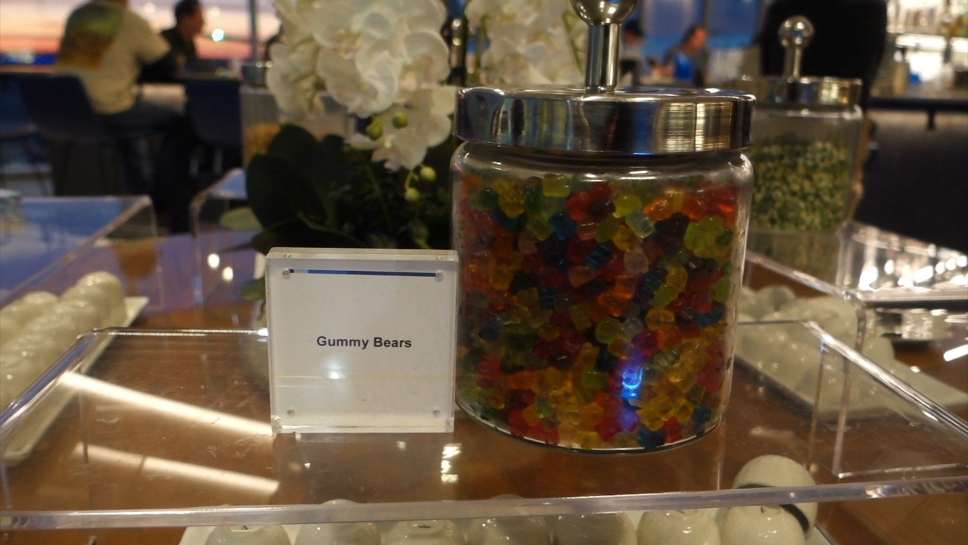 A clear apothecary jar of multicolored gummy bears at the United Club at ORD Concourse C with a printed name placard and a bowl of mints in the foreground.