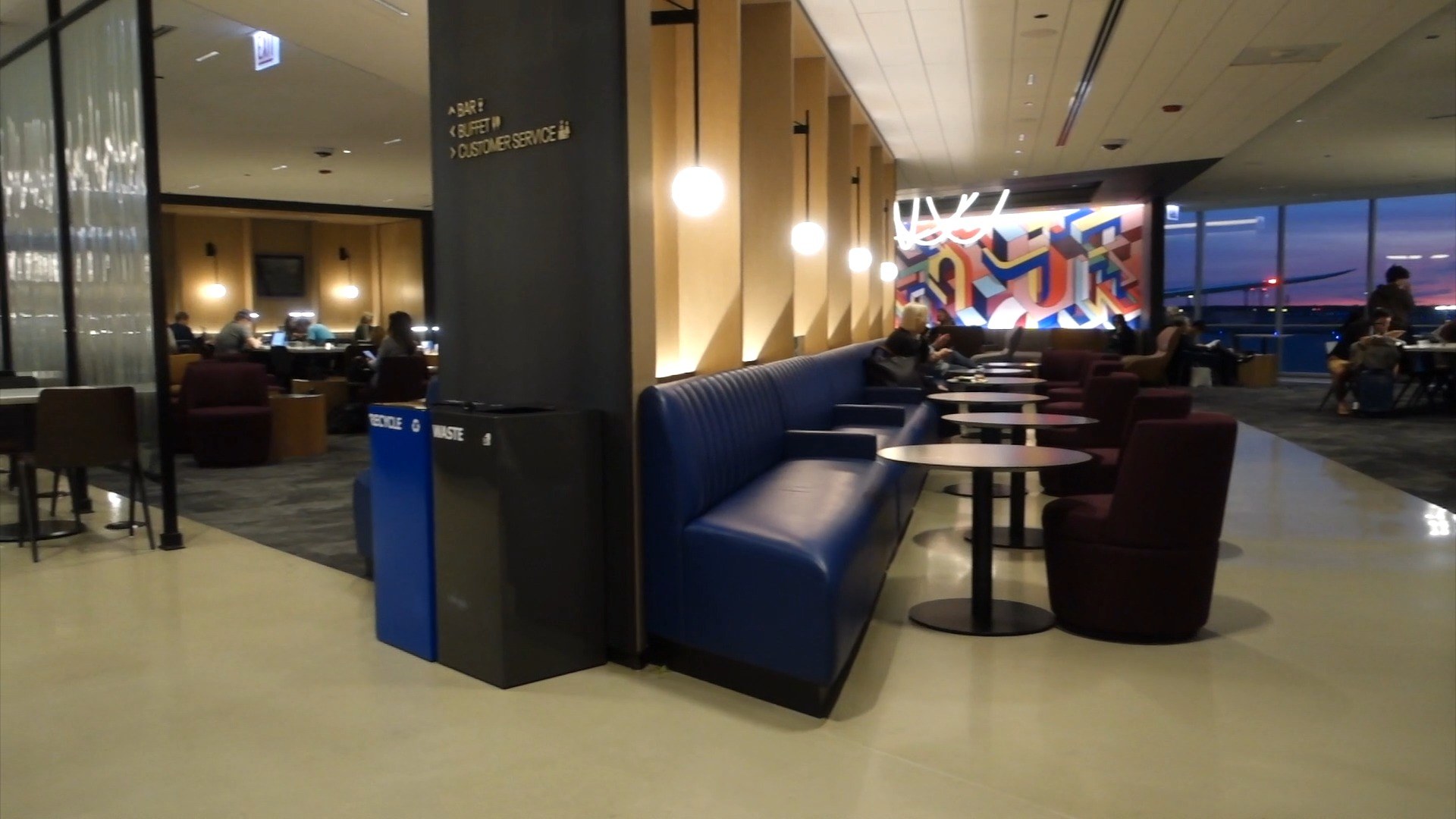Wide view of the United Club at ORD Concourse C with the signature abstract art mural, neon ceiling sculpture, blue banquette seating, and the sunset visible through the windows.