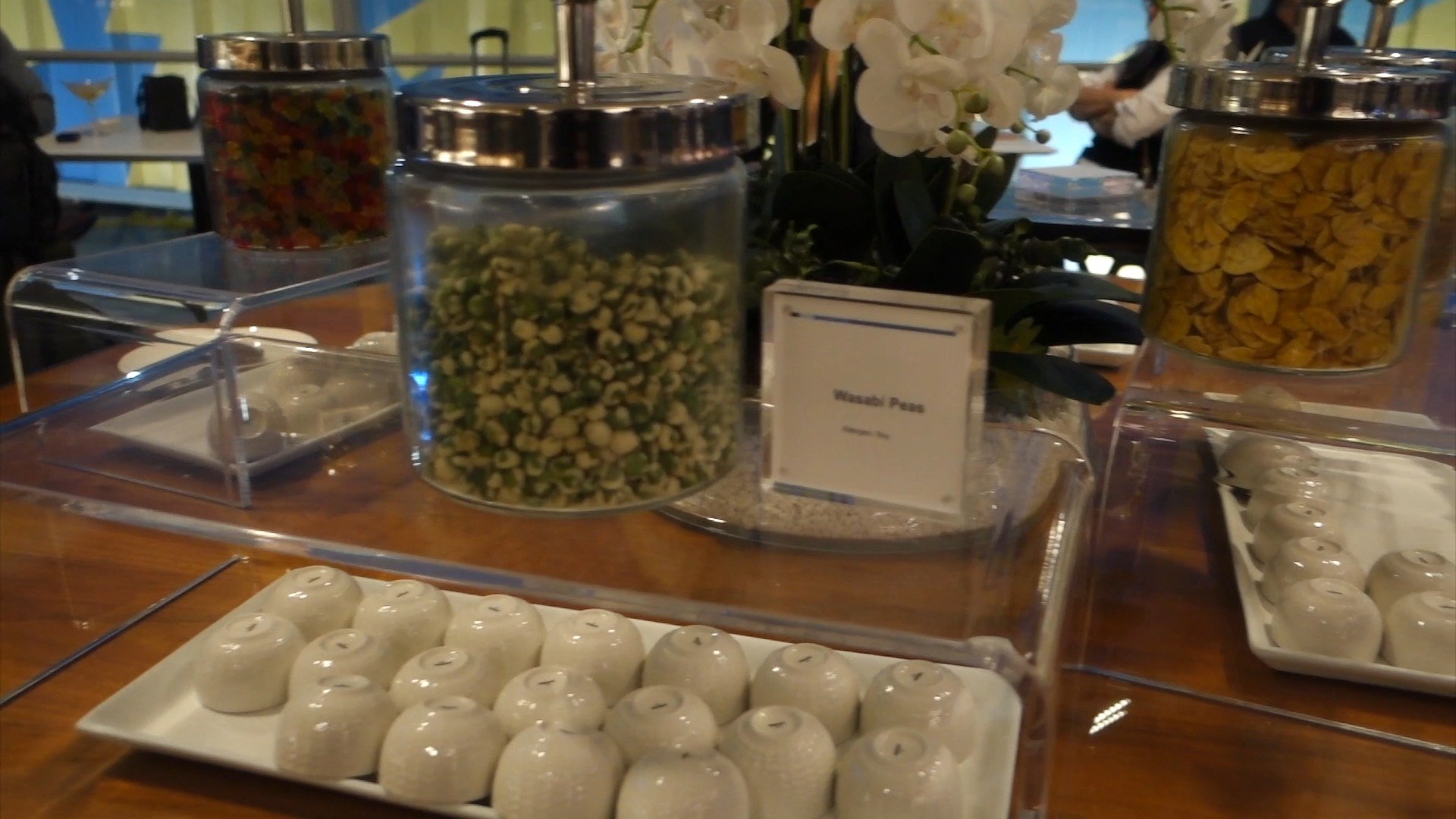 Glass apothecary jars of wasabi peas, gummy worms, and corn nuts on the snack counter at the United Club at ORD Concourse C, with white orchids between them.