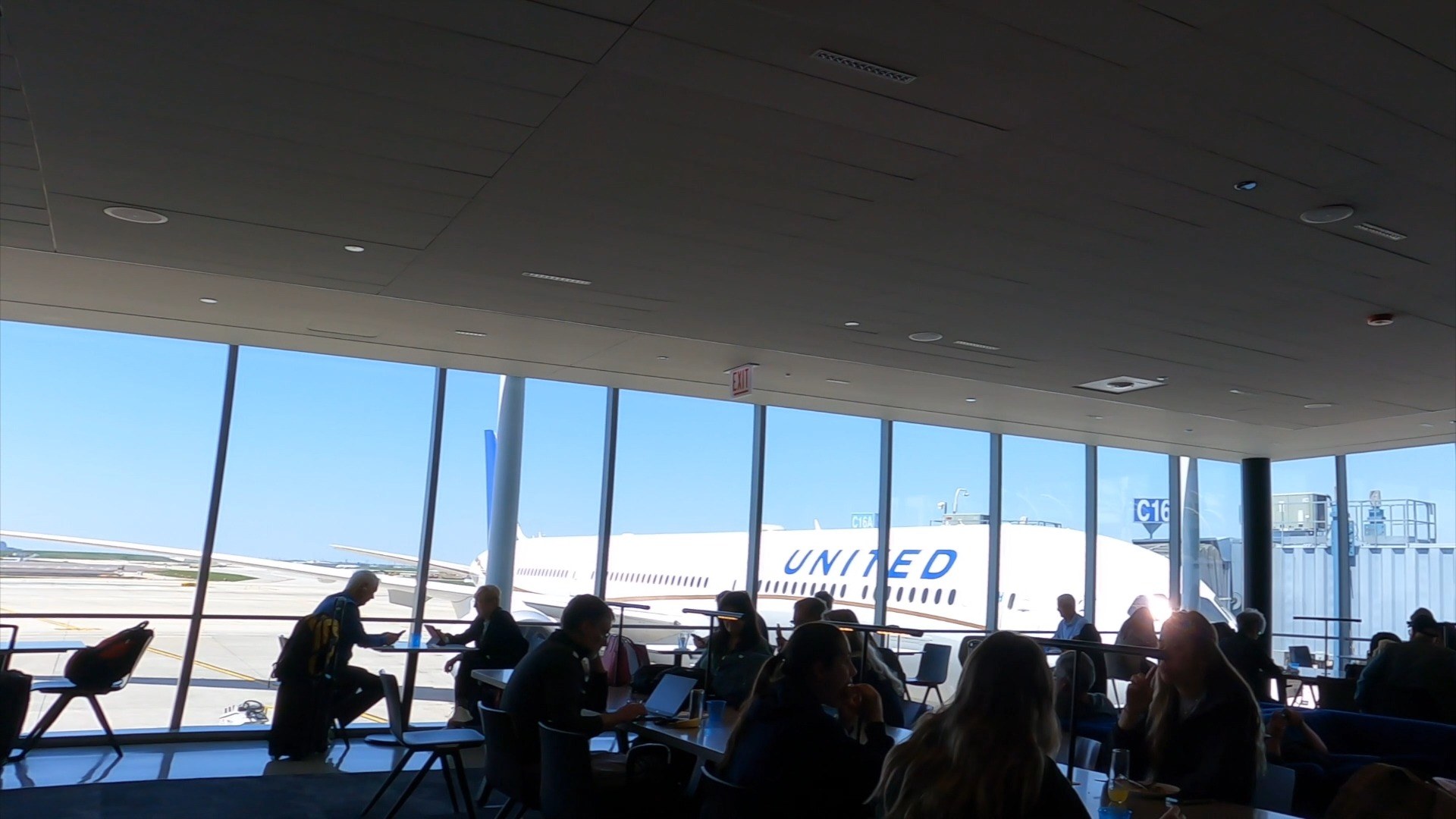 Tarmac view from the United Club at ORD Concourse C with a United widebody parked at Gate C16 and travelers working at high-tops along the floor-to-ceiling windows.