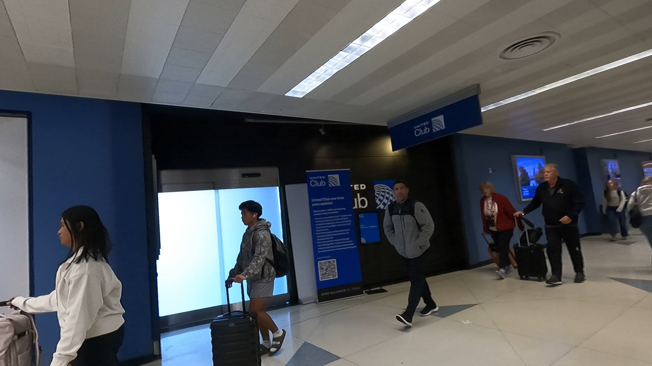 Approach to the United Club at ORD Concourse F with the United Club hanging sign overhead, blue accent walls, and travelers walking down the corridor.