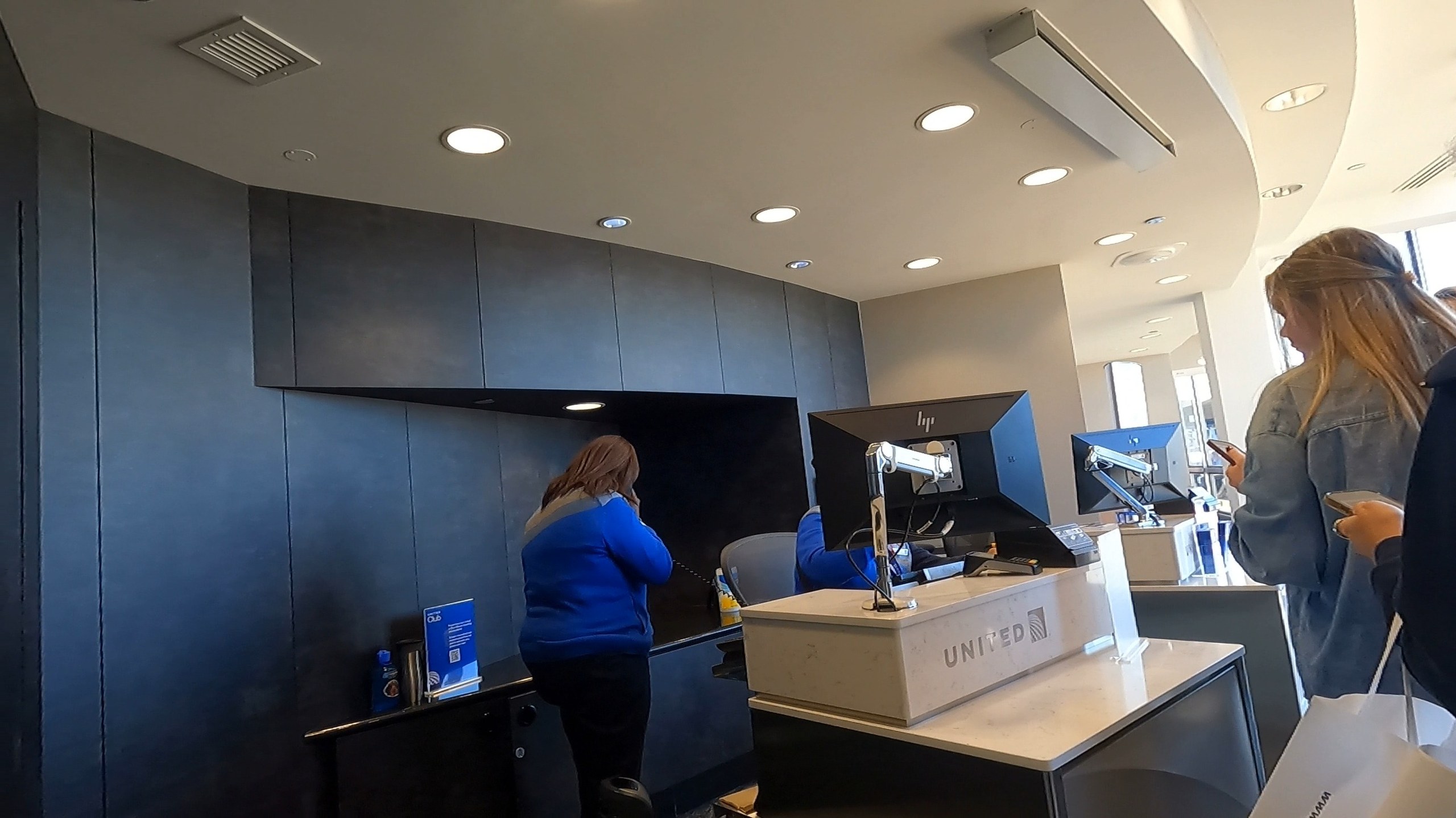Front desk at the United Club at ORD Concourse F with the United wordmark on white marble, dark navy paneling, and an agent on the phone behind dual HP monitors.