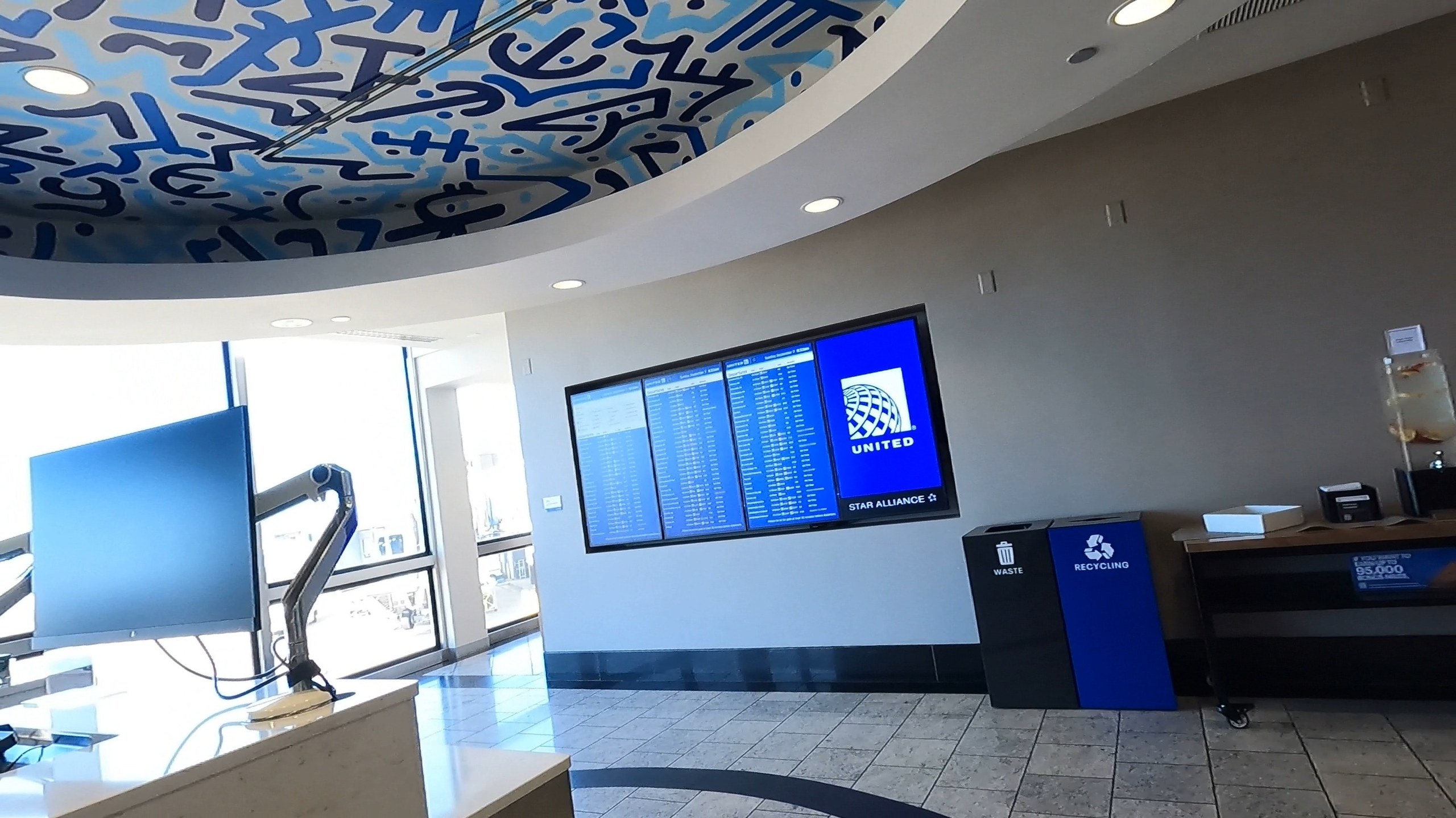 Lobby of the United Club at ORD Concourse F with the signature graffiti-art ceiling overhead, a flight-information screen on the wall, and recycling bins on the right.