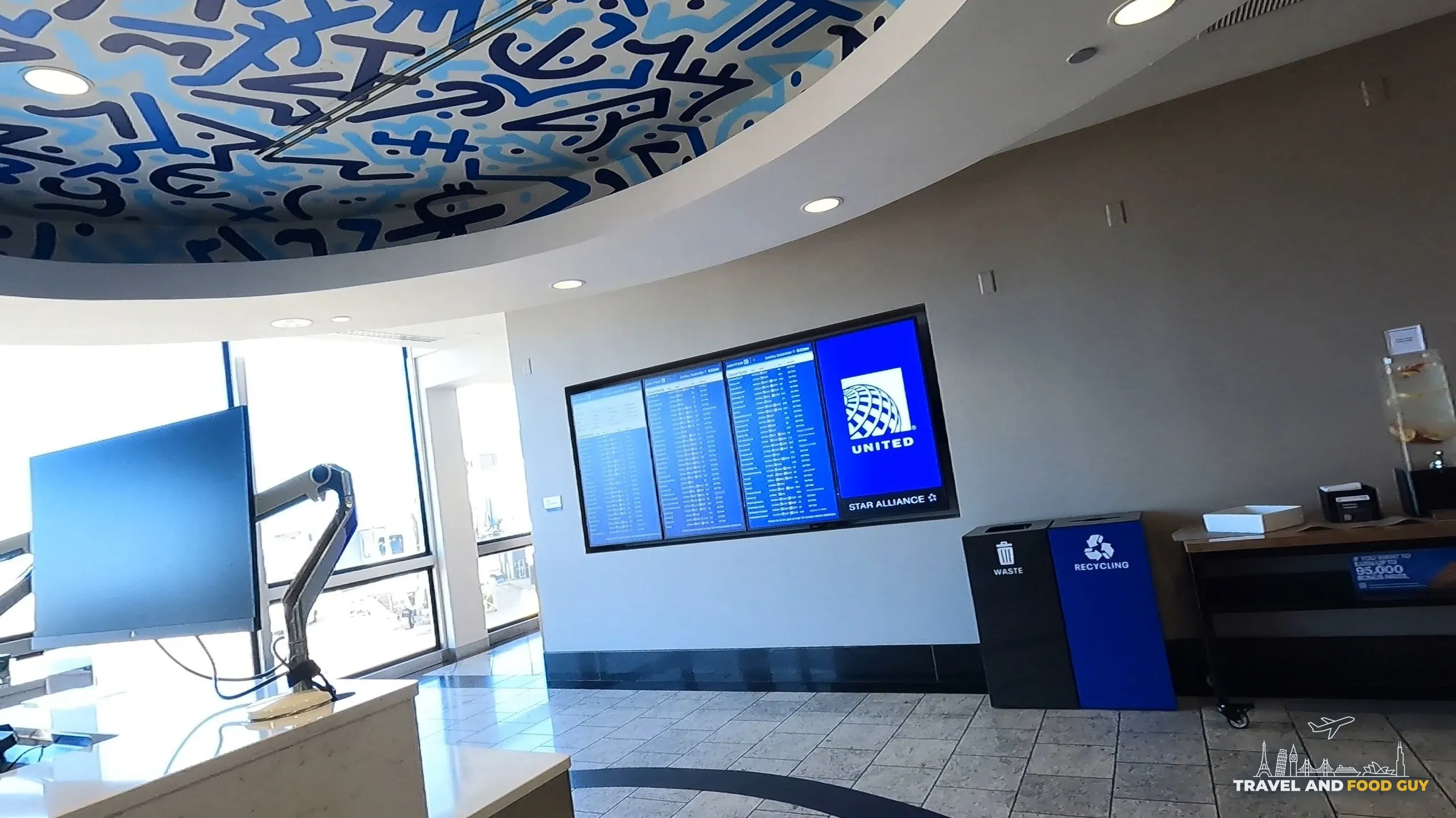 Lobby of the United Club at ORD Concourse F with the signature graffiti-art ceiling overhead, a flight-information screen on the wall, and recycling bins on the right.