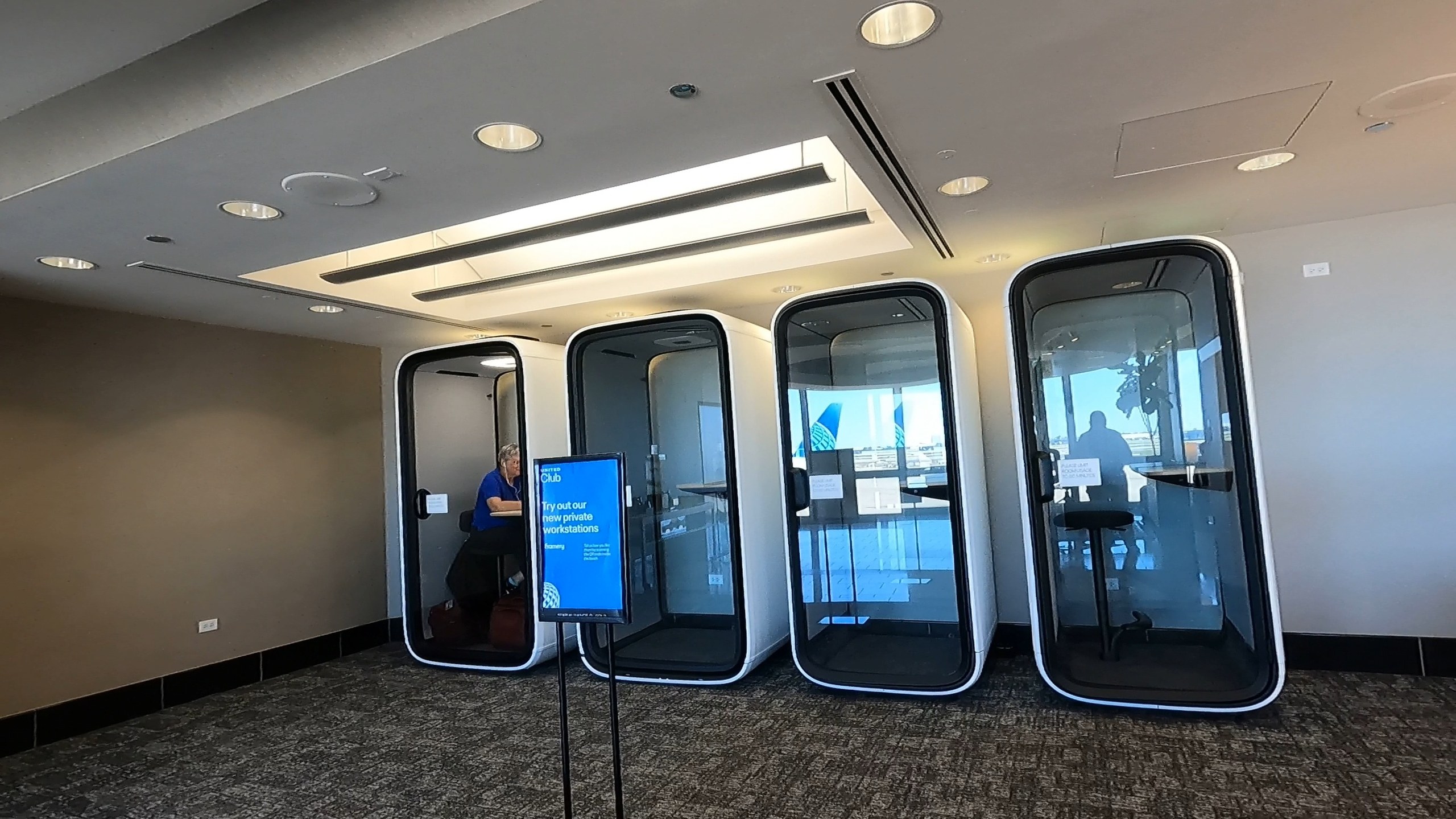 Four modern enclosed phone booth pods at the United Club at ORD Concourse F under a recessed light coffer, with the Try out our newest private workstations sign in front.