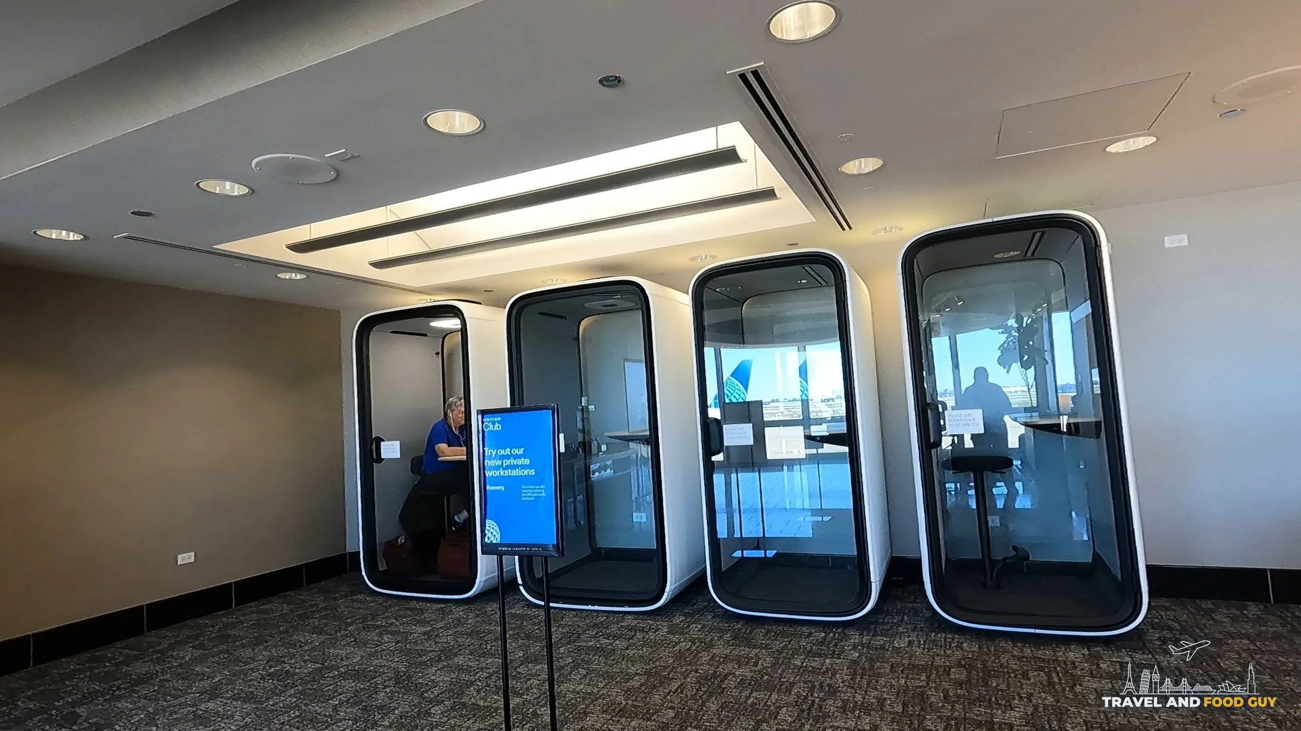 Four modern enclosed phone booth pods at the United Club at ORD Concourse F under a recessed light coffer, with the Try out our newest private workstations sign in front.