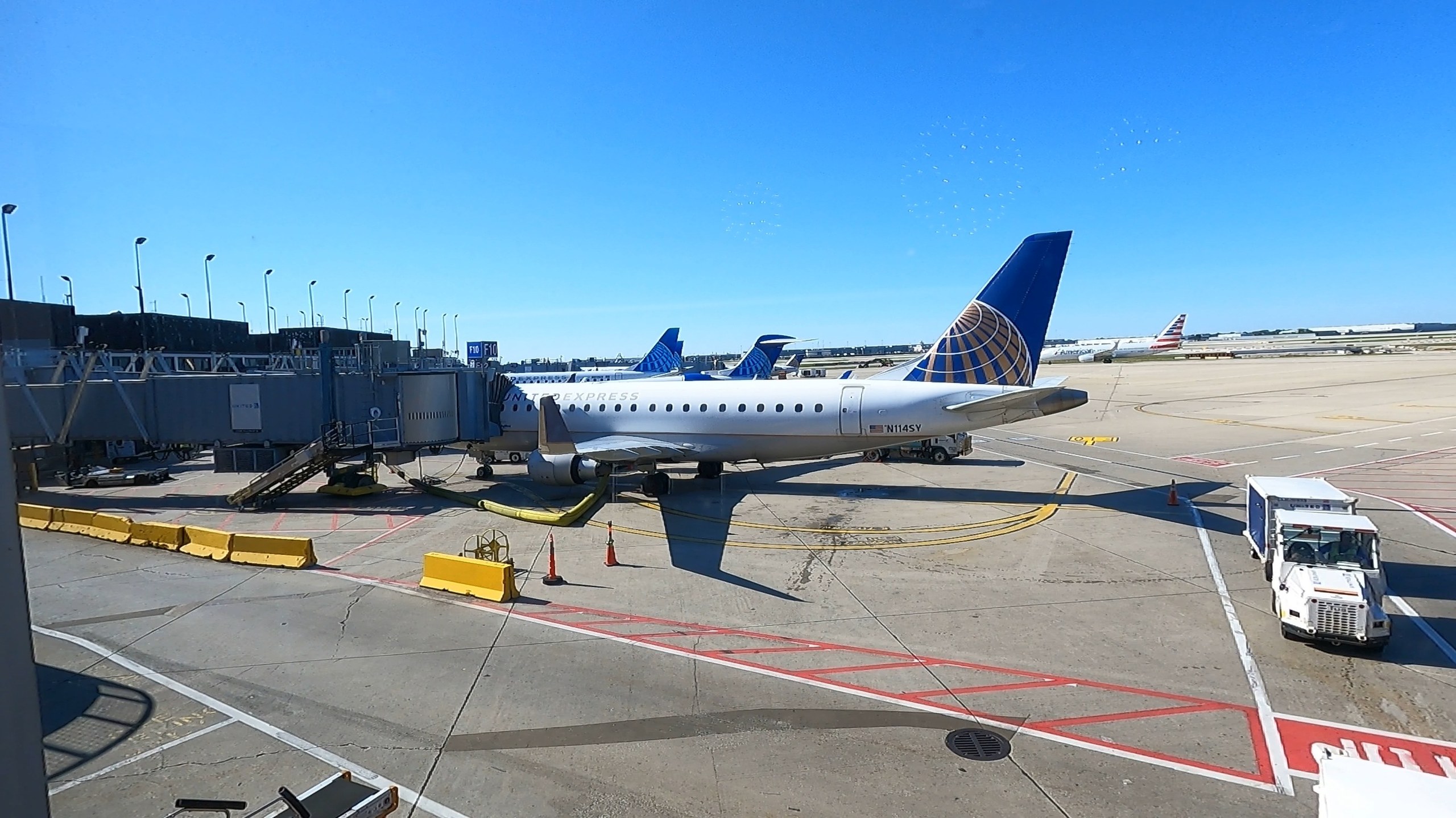 Tarmac view from the United Club at ORD Concourse F with a United Express Embraer E175 regional jet at the gate and a clear blue sky overhead.