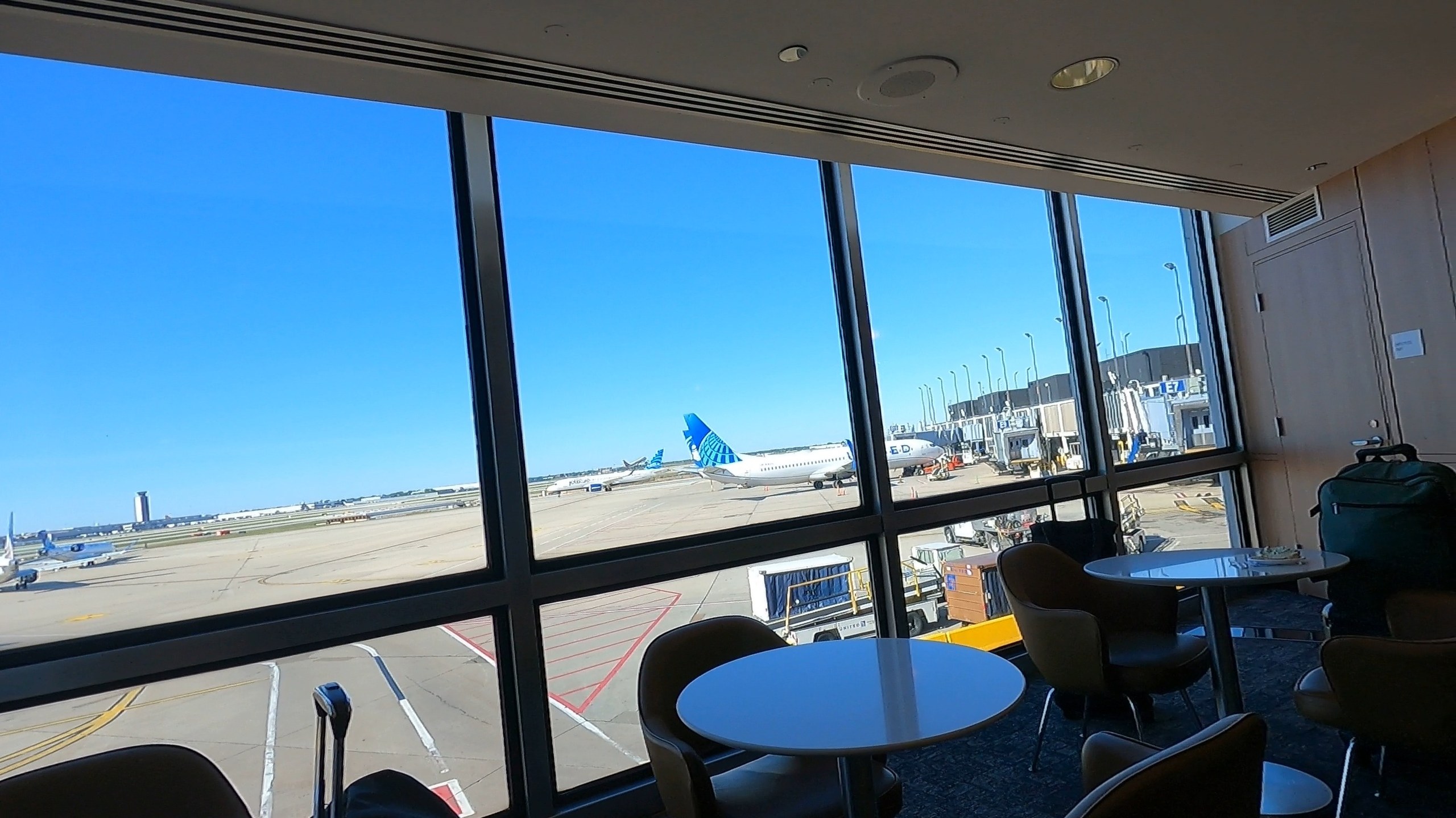 Floor-to-ceiling tarmac view at the United Club at ORD Concourse F with a United plane visible at the gate and small round tables in the foreground.