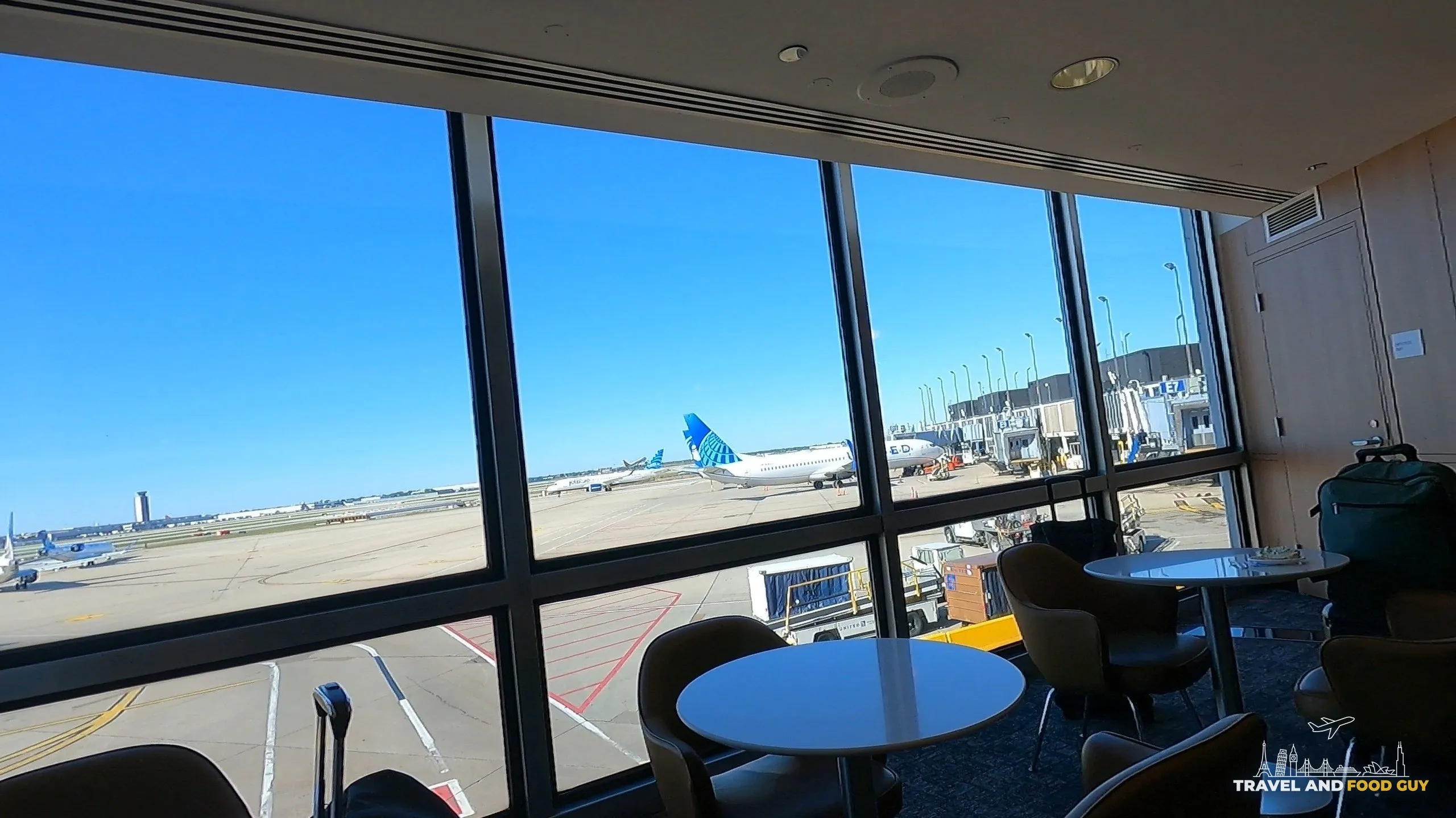 Floor-to-ceiling tarmac view at the United Club at ORD Concourse F with a United plane visible at the gate and small round tables in the foreground.