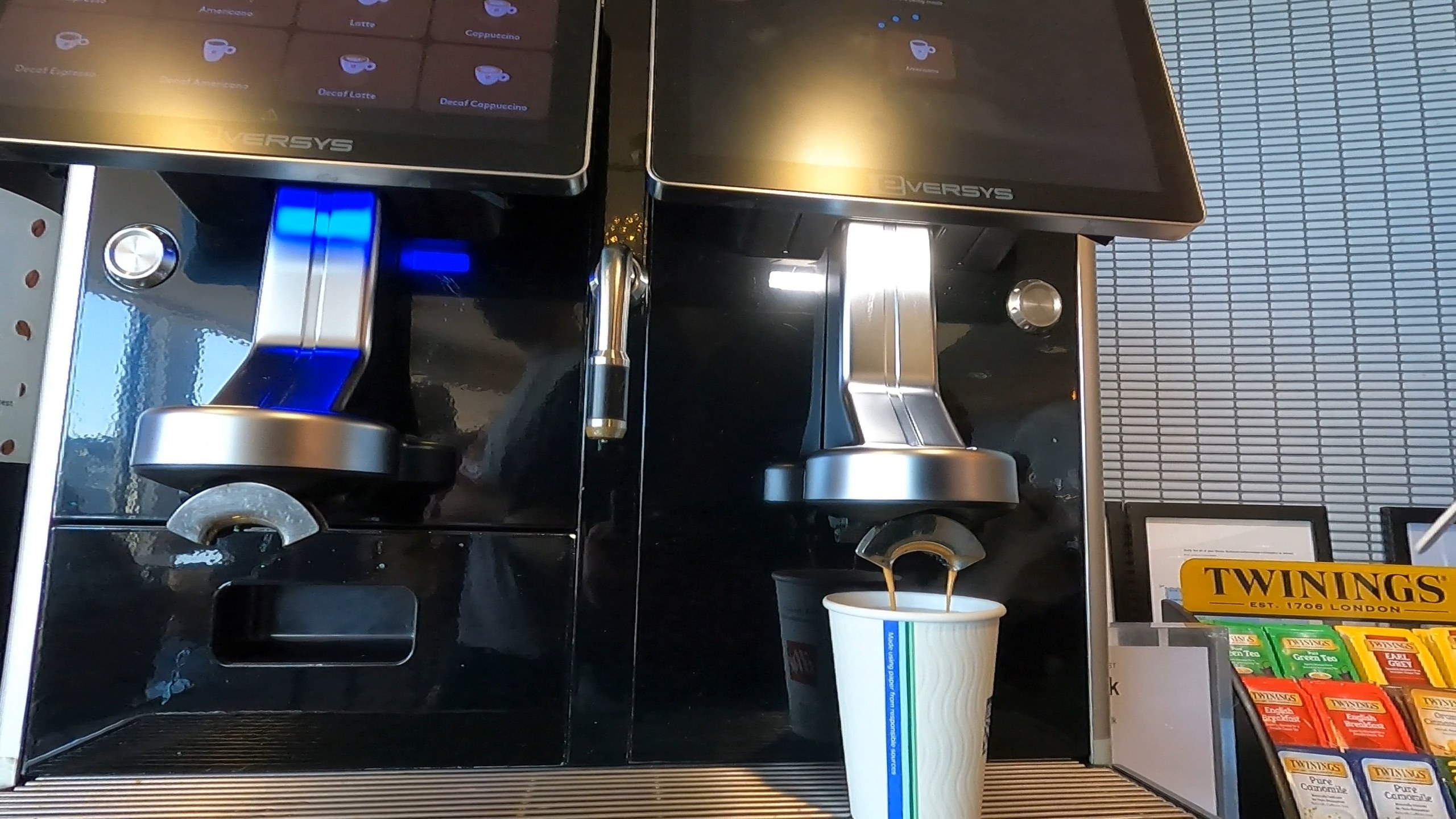 Twin Eversys automatic espresso machines pouring coffee into a paper cup at the United Club at ORD Concourse F, with Twinings tea boxes alongside.