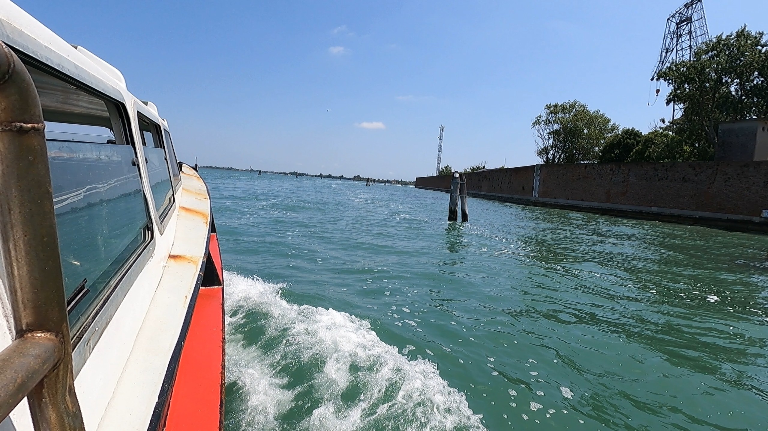 View from the side of a Venice vaporetto crossing the lagoon with open water and a low brick wall in the distance